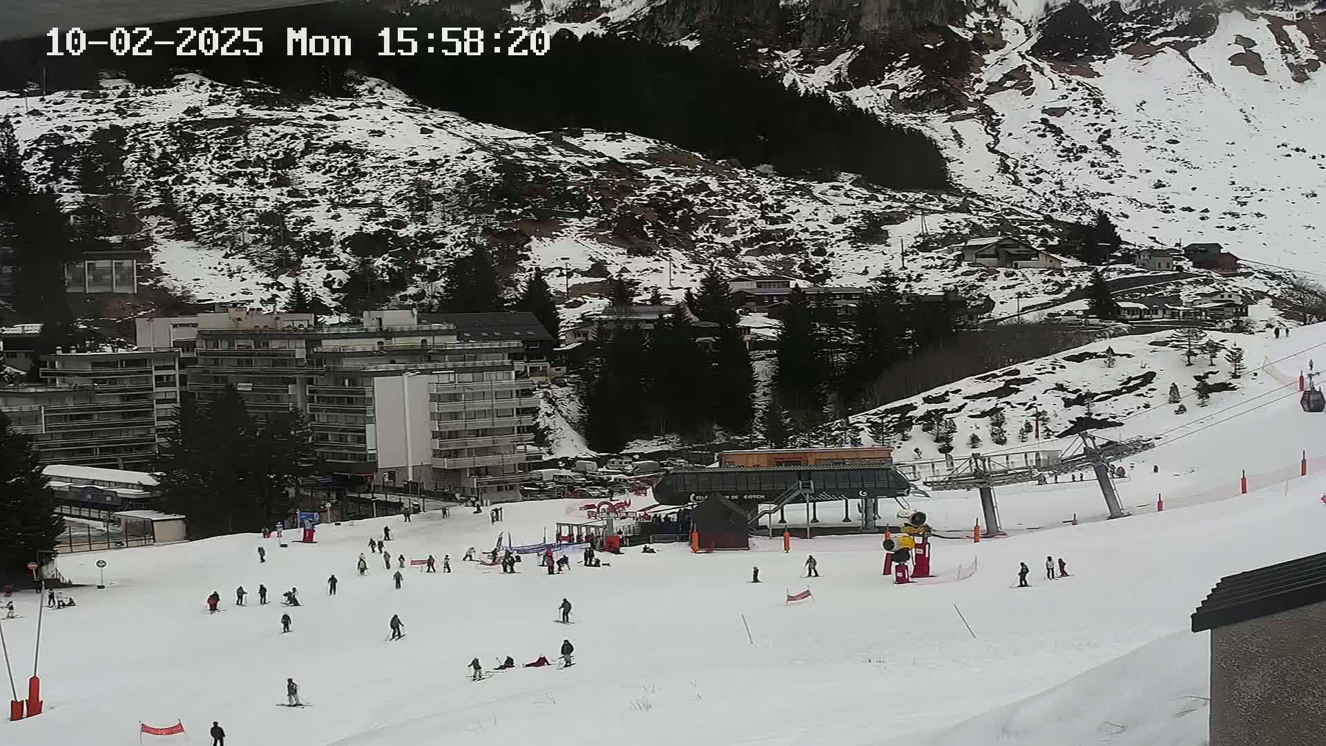 A snowy ski slope with several skiers and a ski lift, situated below snow-covered mountains and buildings on an overcast day.