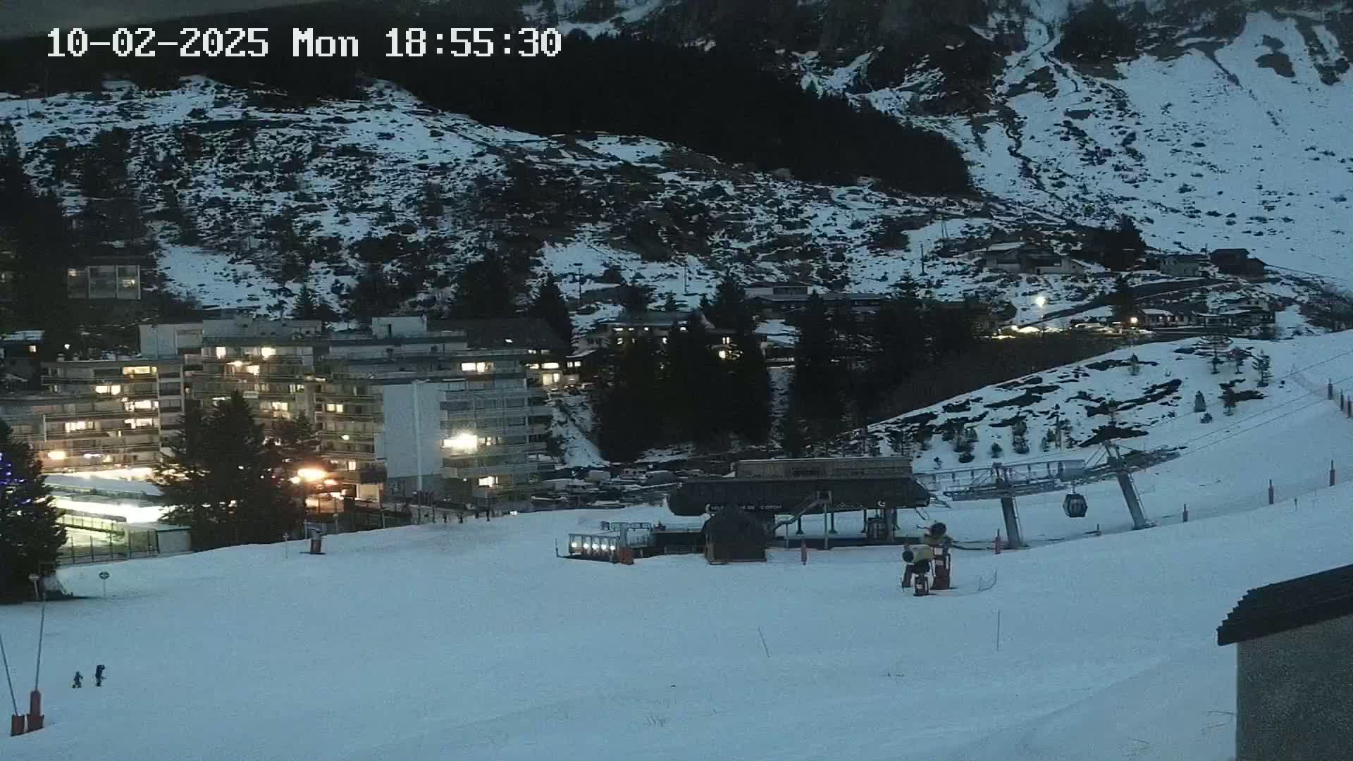 A snow-covered ski slope at twilight, with buildings, a ski lift, and a few people visible.