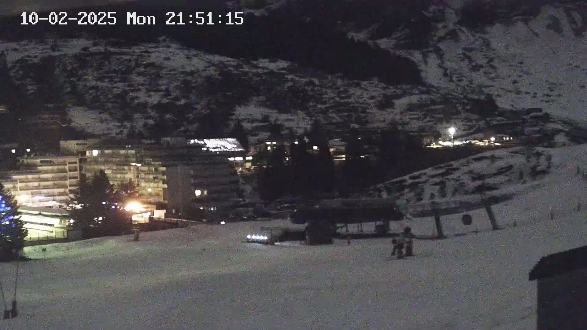 A snow-covered ski slope at night, illuminated by lights from a nearby building and a few scattered lights, shows a few people and a snowcat.