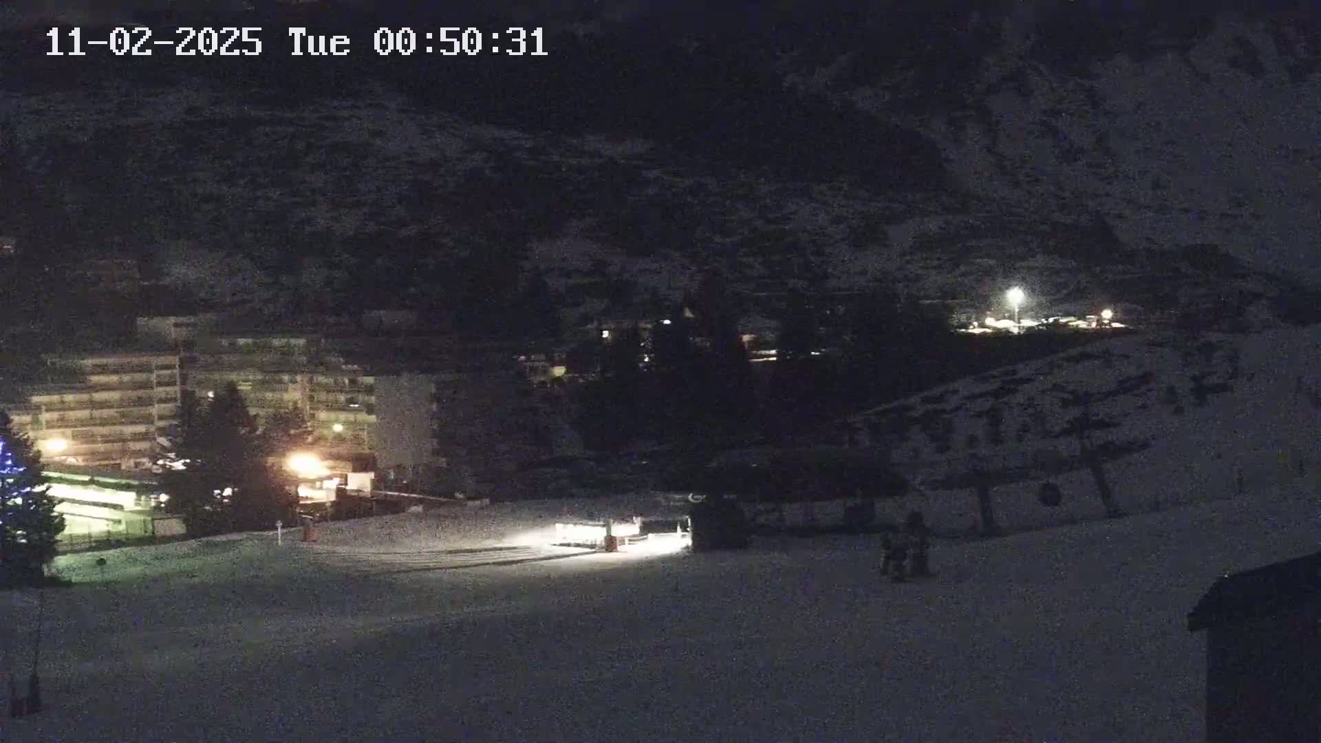 A nighttime, snow-covered ski slope is shown, with a building and some lights visible in the background.