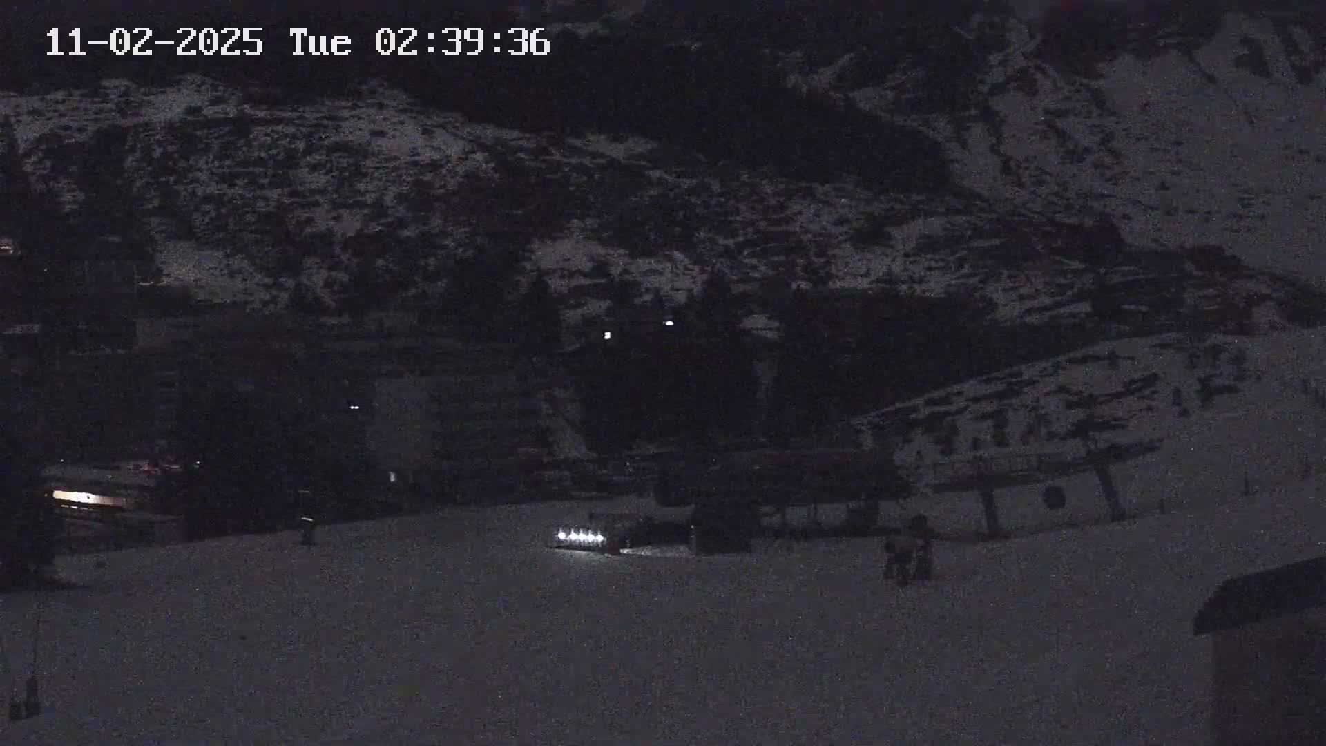 A nighttime, snow-covered landscape shows a few lights and dark shapes suggesting buildings and possibly people in the distance.