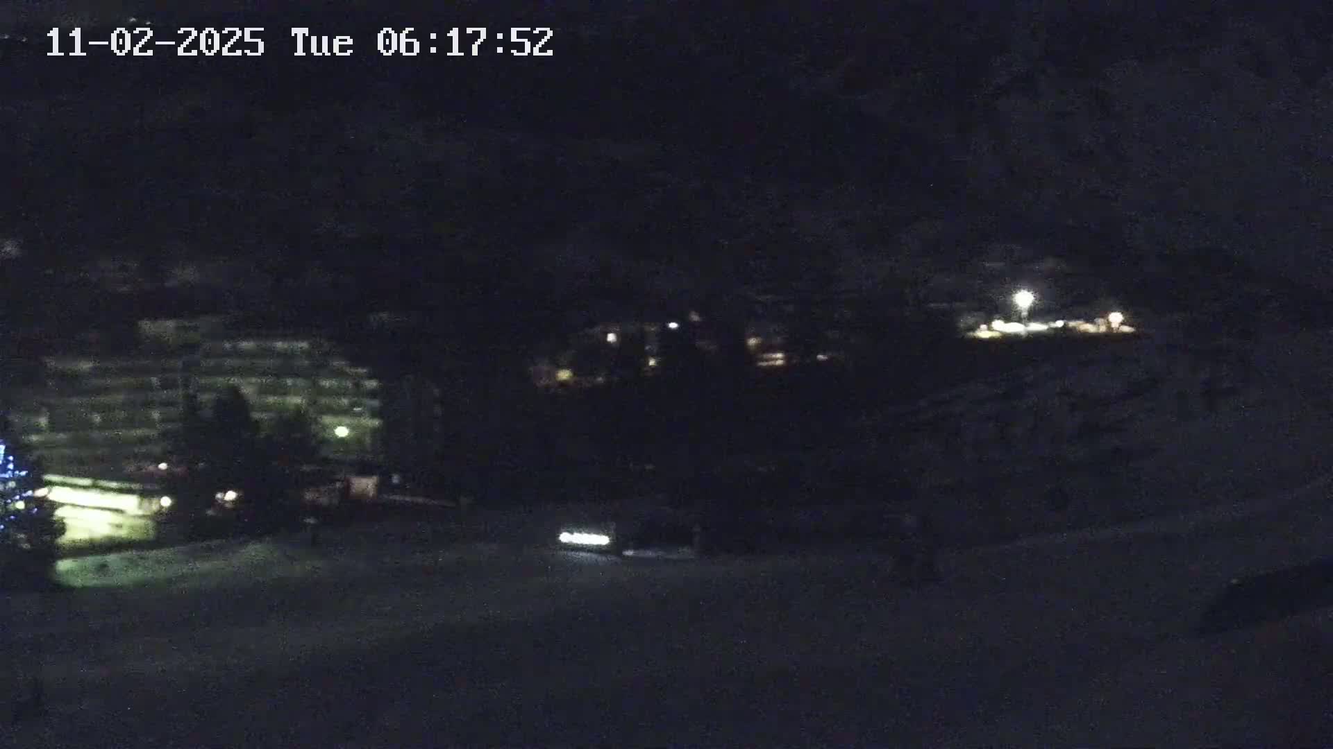 A nighttime, snowy scene shows a large building with lights, a vehicle with its headlights on, and some distant lights in the background.