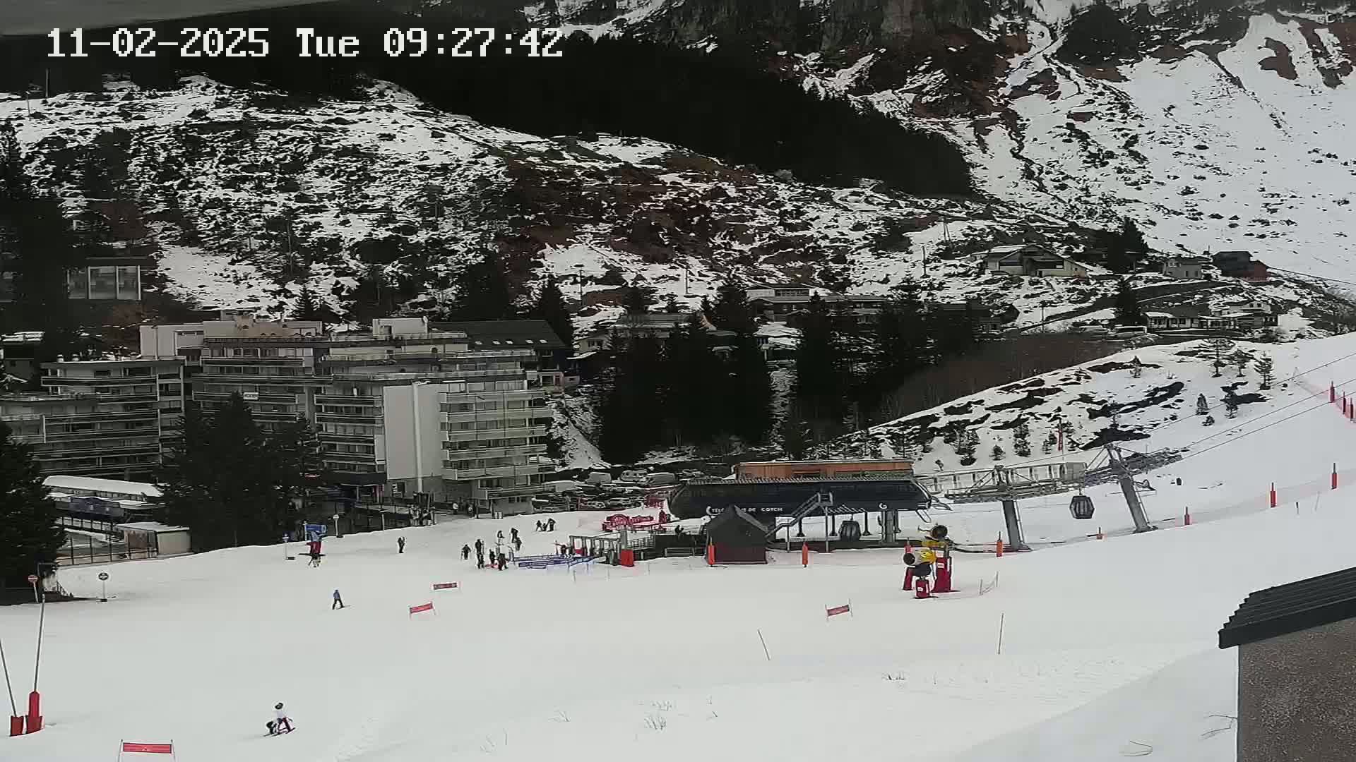 A snowy ski slope with a chairlift, several buildings, and a few skiers visible on a partly cloudy day.