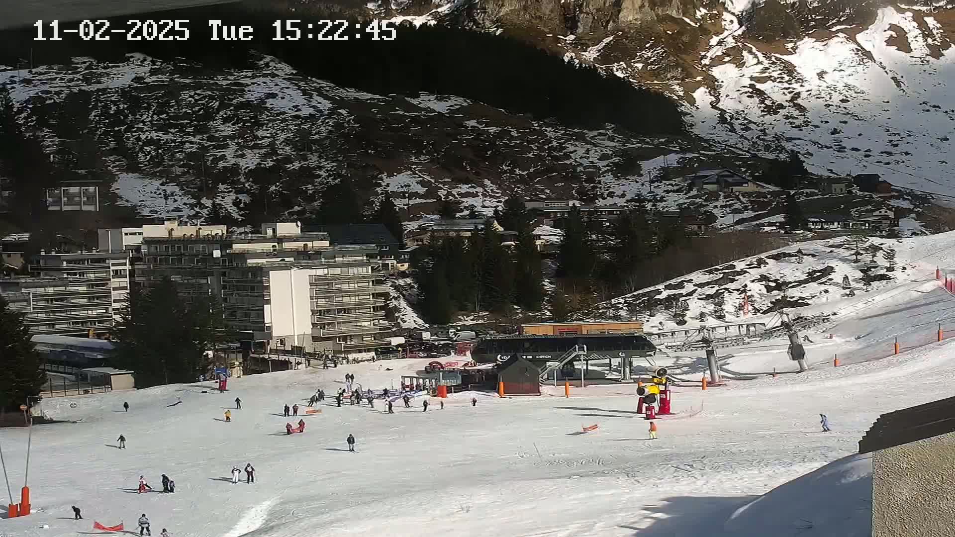 A snowy ski slope with a ski lift, several buildings, and scattered skiers and snowboarders under a sunny sky.