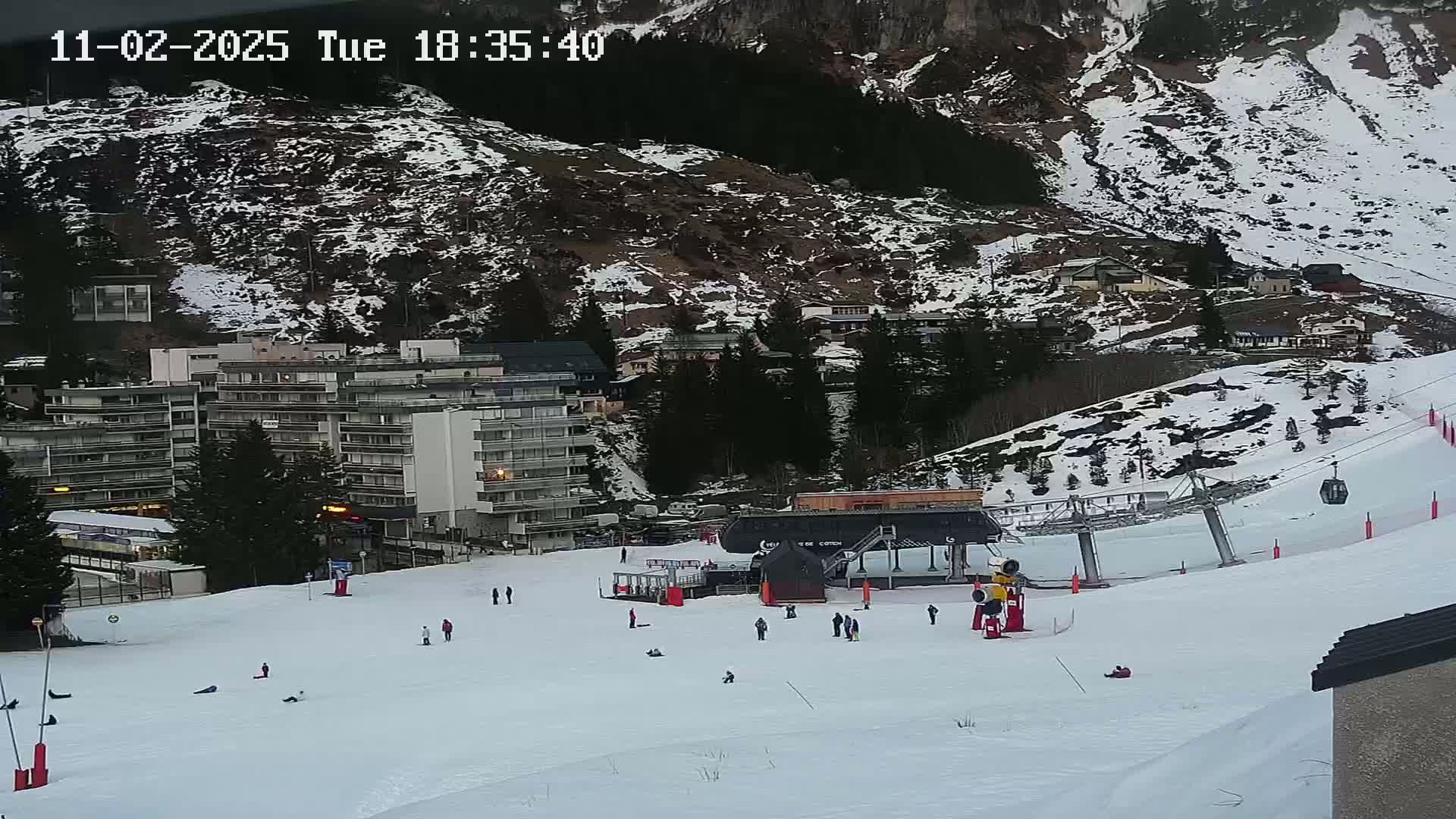 A snow-covered ski slope with a chairlift and several people skiing or snowboarding, situated below a mountainside with buildings and some patchy snow.