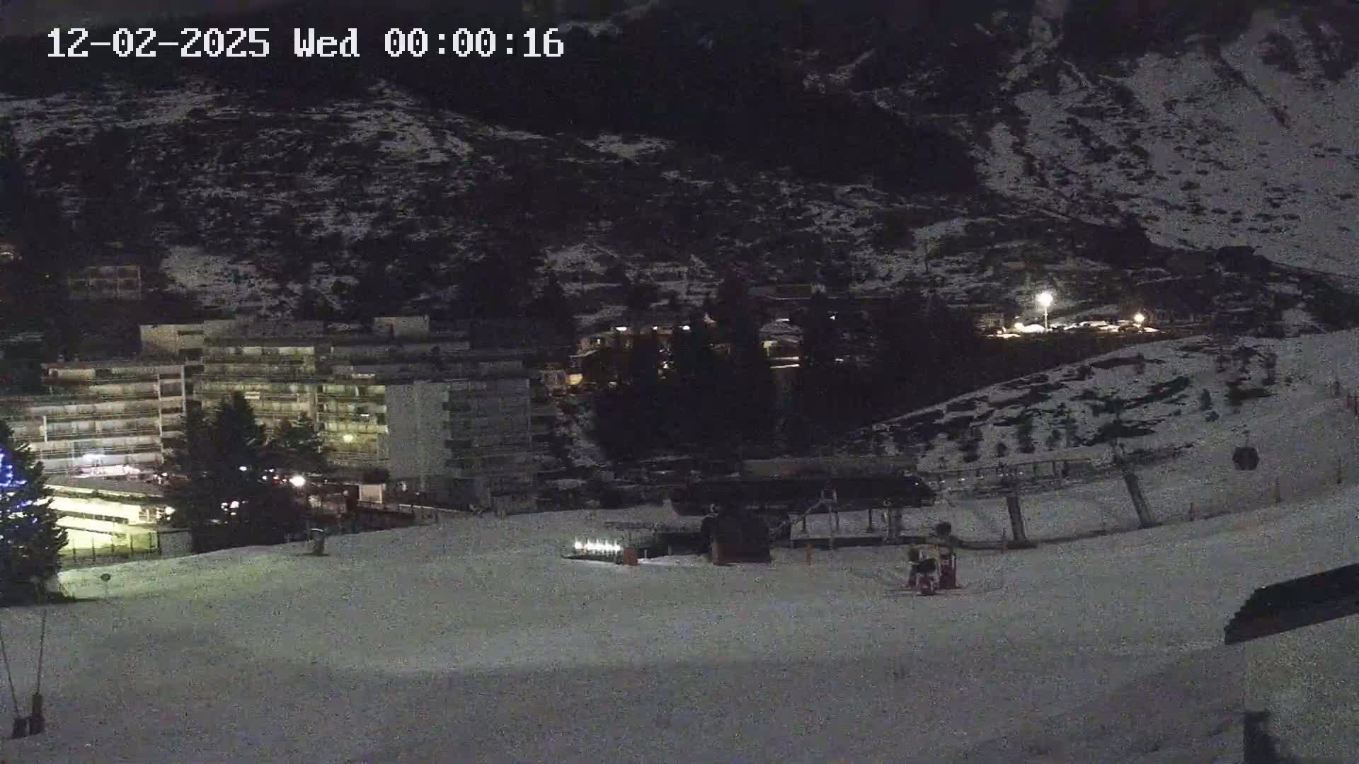 A snow-covered ski slope at night, with a multi-story building and sparsely lit buildings in the background under dark skies.