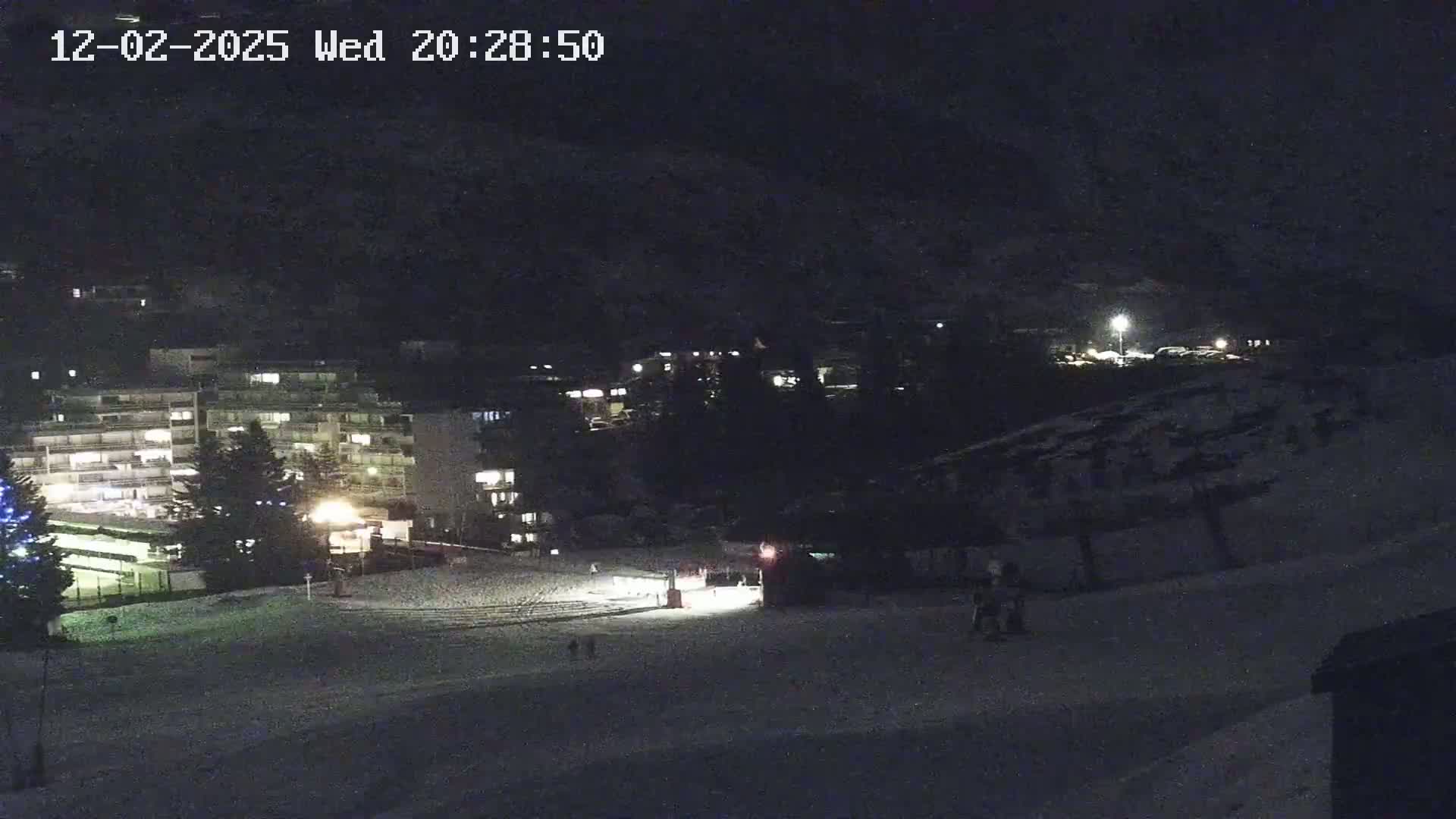 A nighttime view of a snow-covered area with buildings and a few people visible under a dark sky.