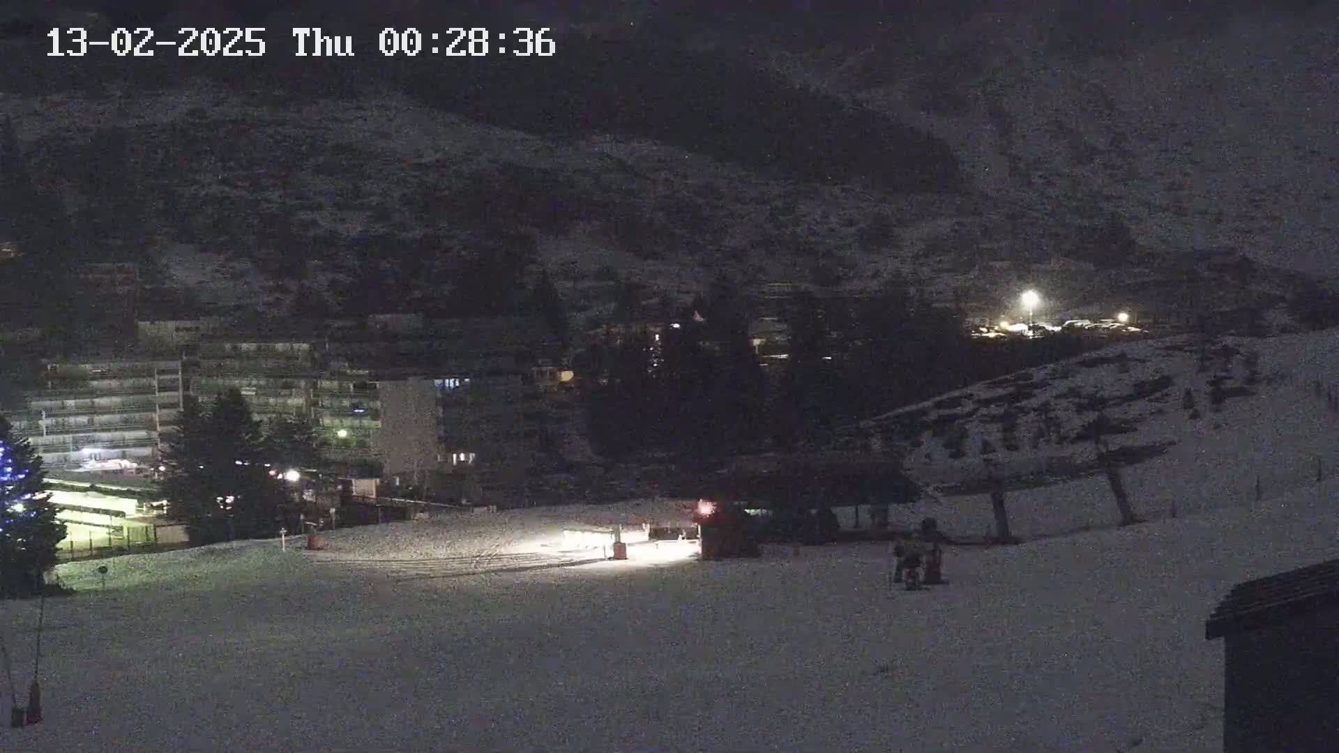 A snow-covered ski slope at night, with a few people visible near a lift, and buildings and a hill in the background under dark, nighttime conditions.