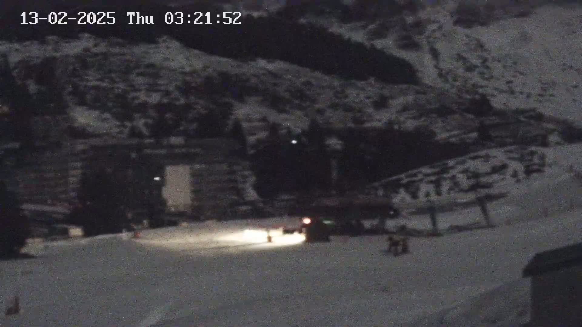 A snow-covered landscape at night, illuminated by headlights of a vehicle near a building and a few people.