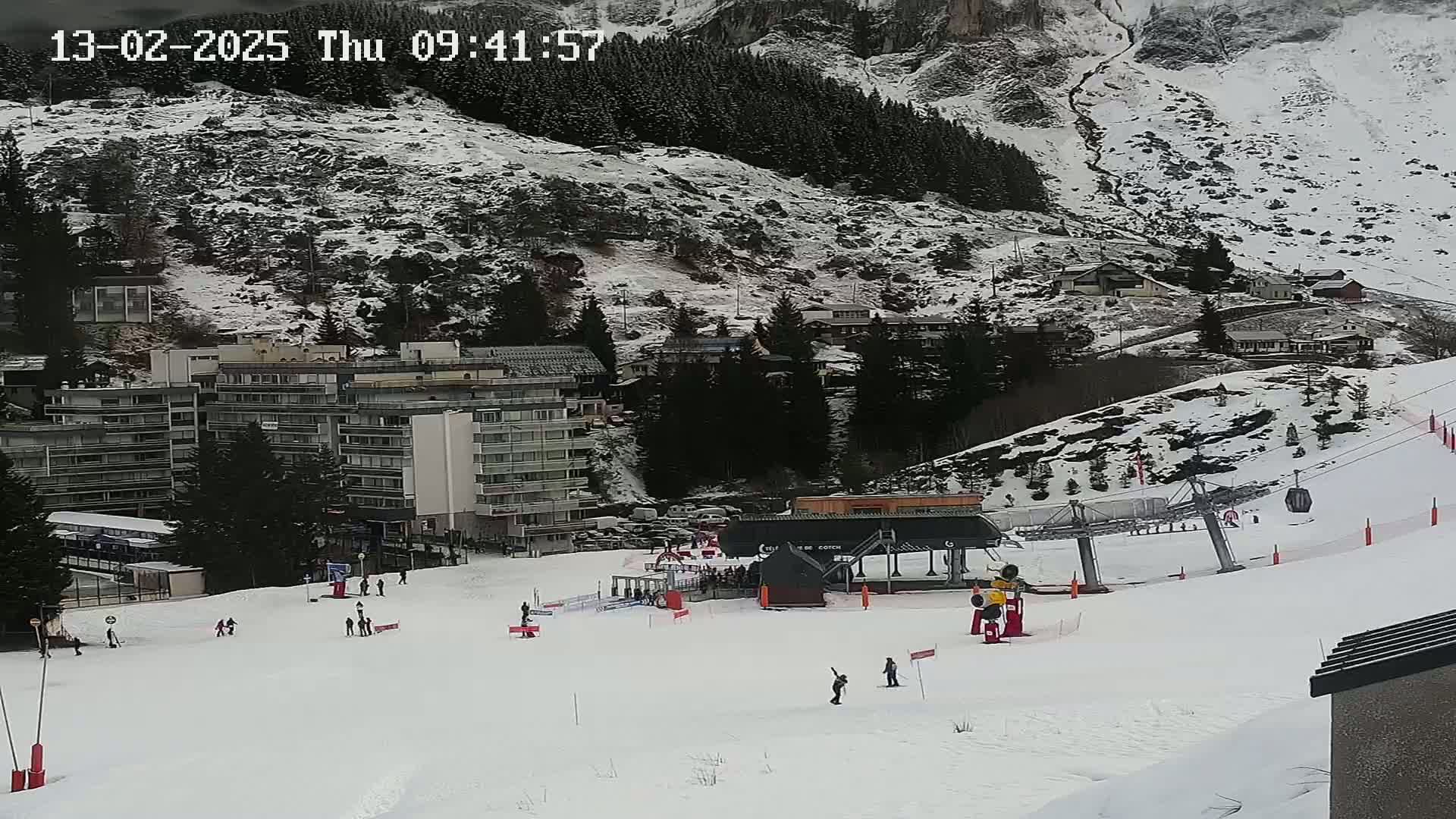 A snowy ski slope with a ski lift, several skiers, and a building complex in the background on a cloudy day.