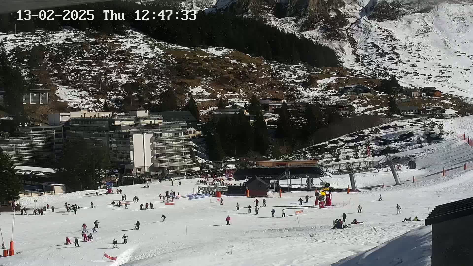 A snowy ski slope with several skiers and snowboarders, a ski lift, and buildings in the background under sunny conditions.