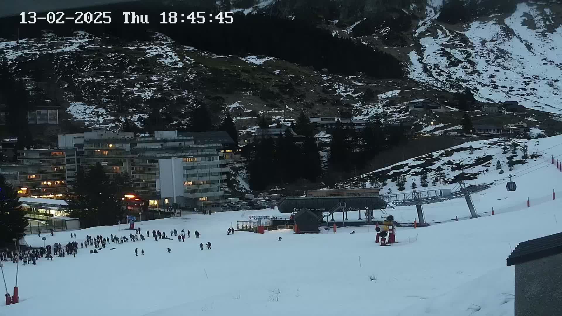 A snow-covered ski slope at dusk, with a ski lift, several buildings with lights on, and a number of people gathered near the bottom of the slope.
