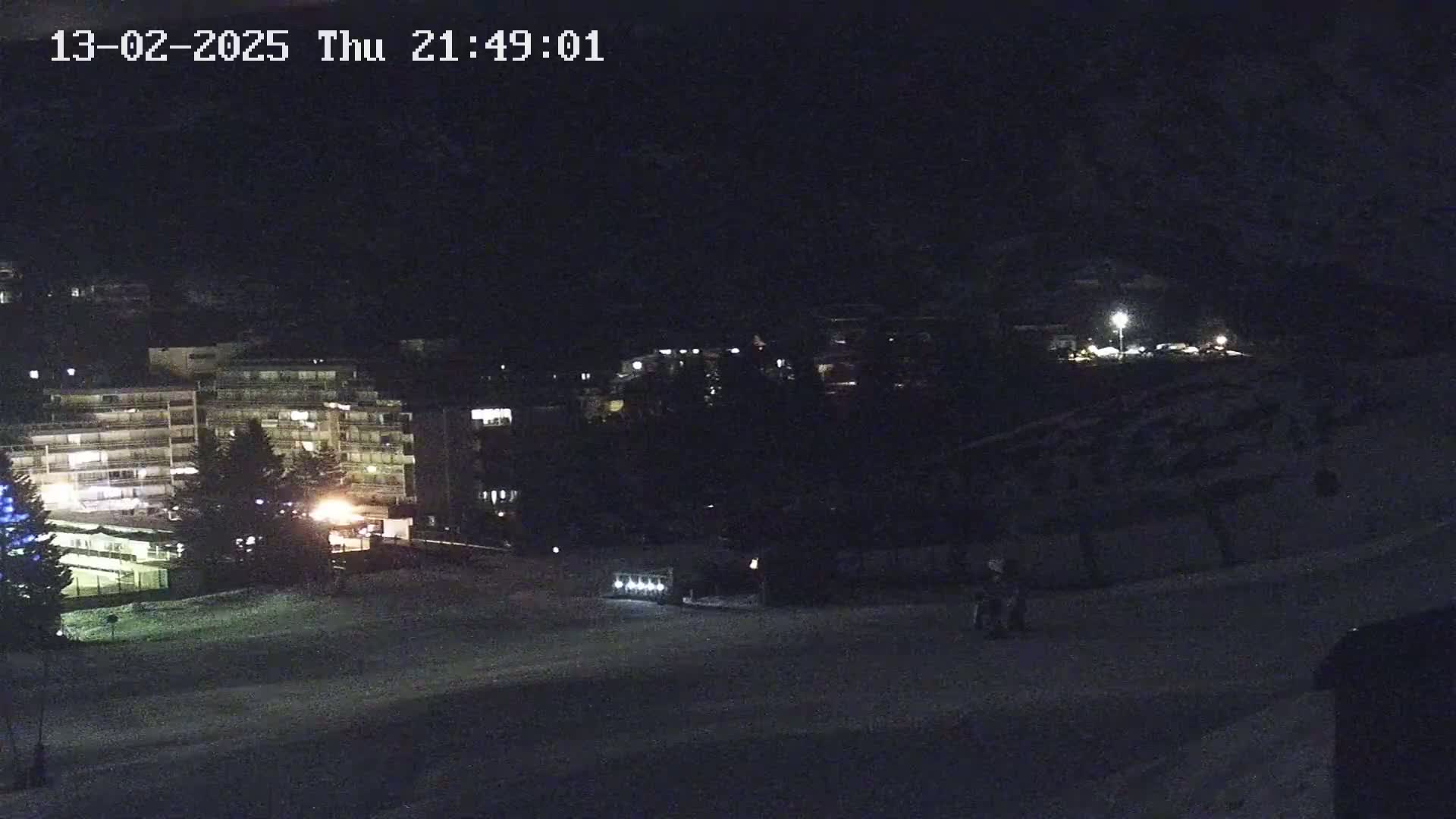 A nighttime view of a snow-covered area with buildings and a few people visible under dark, clear skies.