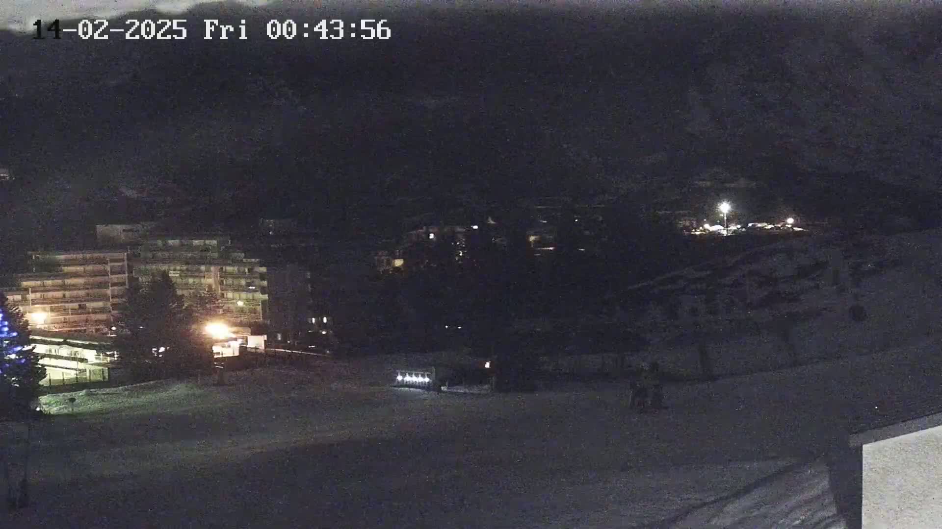 A nighttime, snow-covered scene shows a few buildings with lights on, a vehicle, and some figures in the distance.