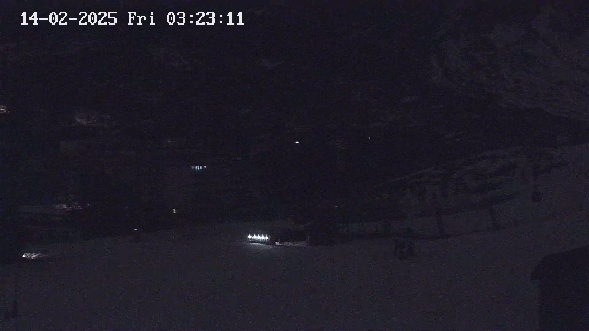 A nighttime, snow-covered scene shows faintly lit objects in the distance under a dark sky.