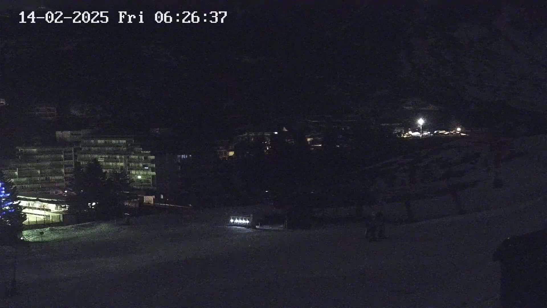 A nighttime view of a snow-covered area with buildings and sparsely lit areas, possibly a ski resort.