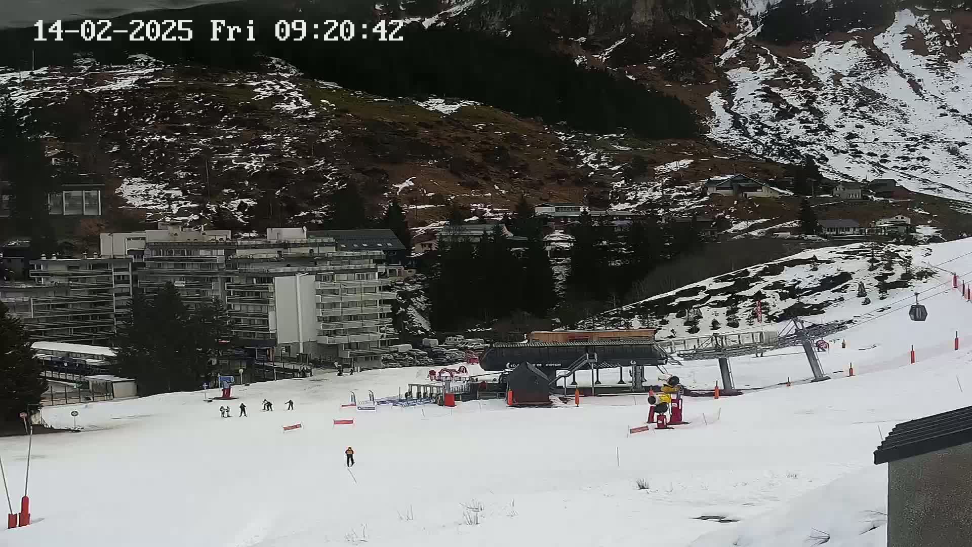 A snow-covered ski slope with a ski lift, several buildings, and a few skiers is seen on a partly cloudy day.