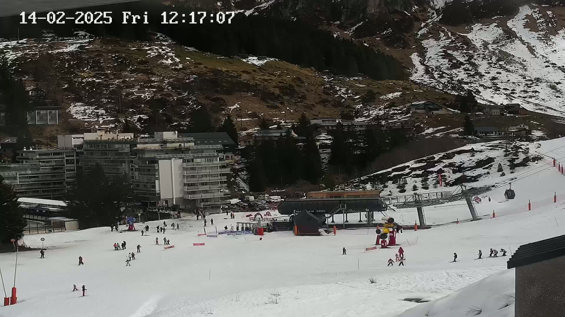 A snowy ski slope with several skiers and a ski lift, situated near a multi-story building and mountainside with patches of snow and bare ground.