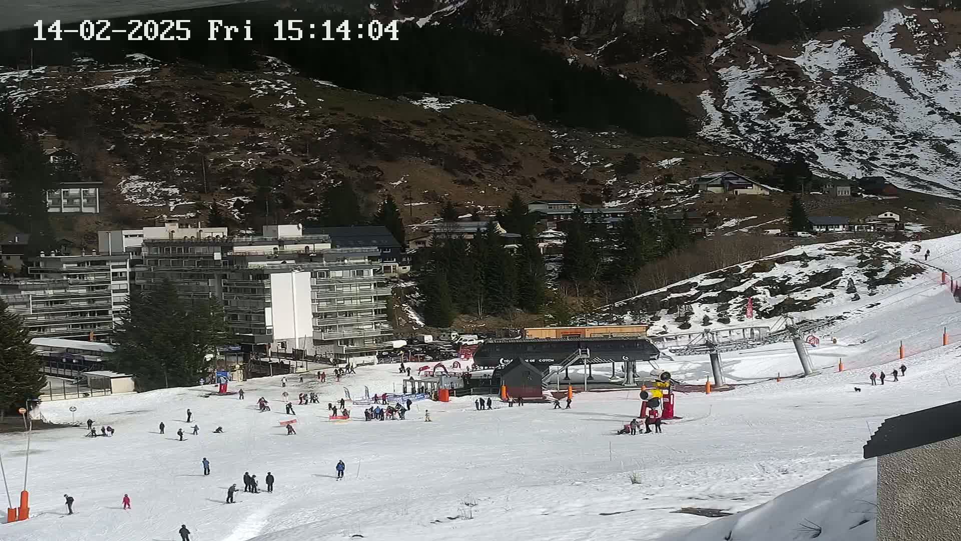 A sunny ski slope with several skiers and snowboarders, a ski lift, and a large apartment building in the background.