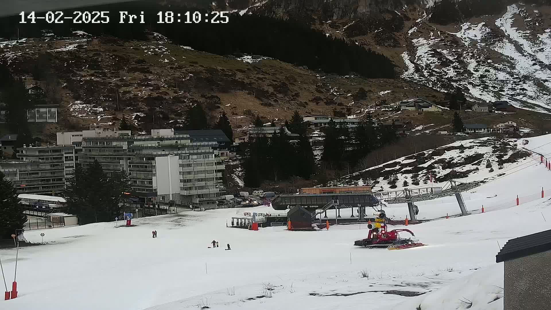 A snow-covered ski slope with a ski lift, a snow groomer, and several buildings in the background on a partly cloudy day.