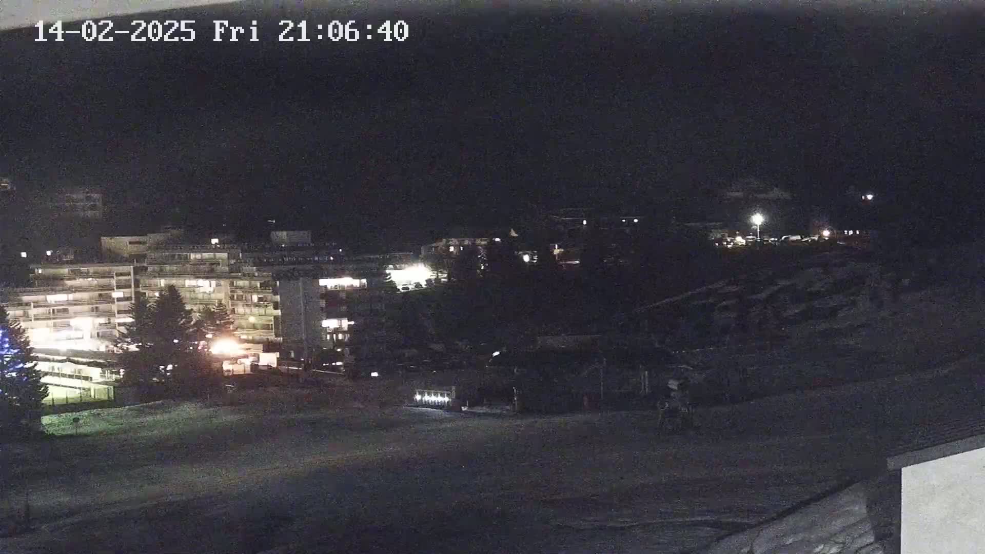 A nighttime, snow-covered scene shows several illuminated buildings and trees, with a few faint lights visible in the distance.
