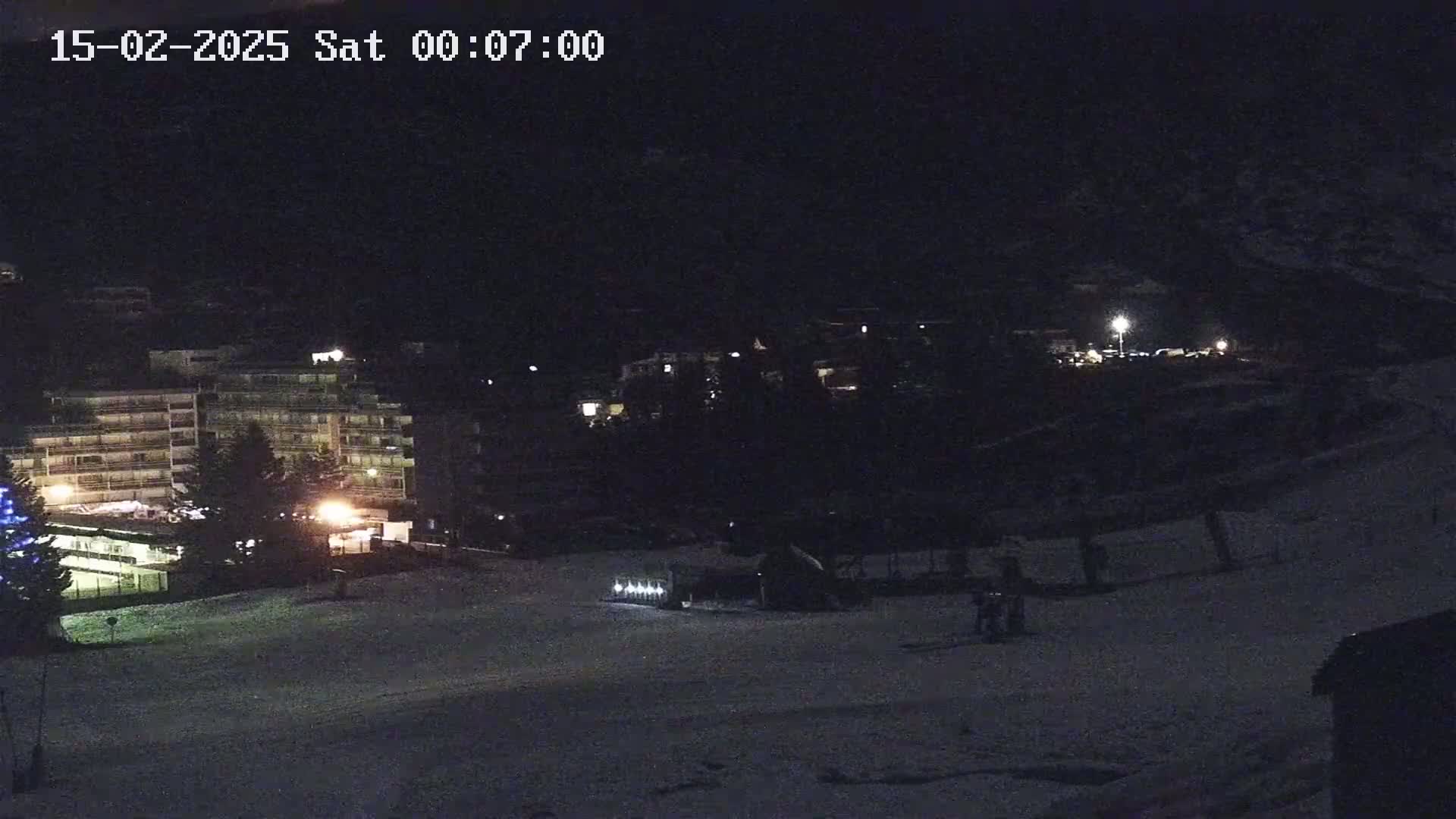A nighttime, snow-covered slope with a building and snow grooming machine visible under dark conditions.