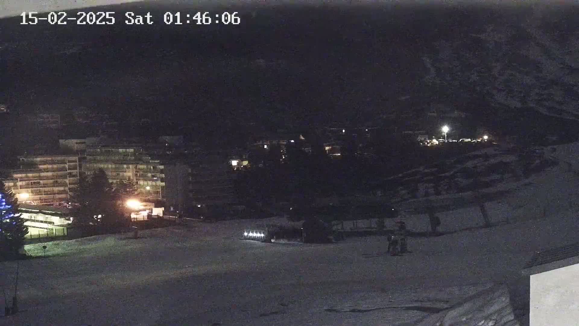 A nighttime, snow-covered scene shows a multi-story building and a few people near a snowcat on a slope.