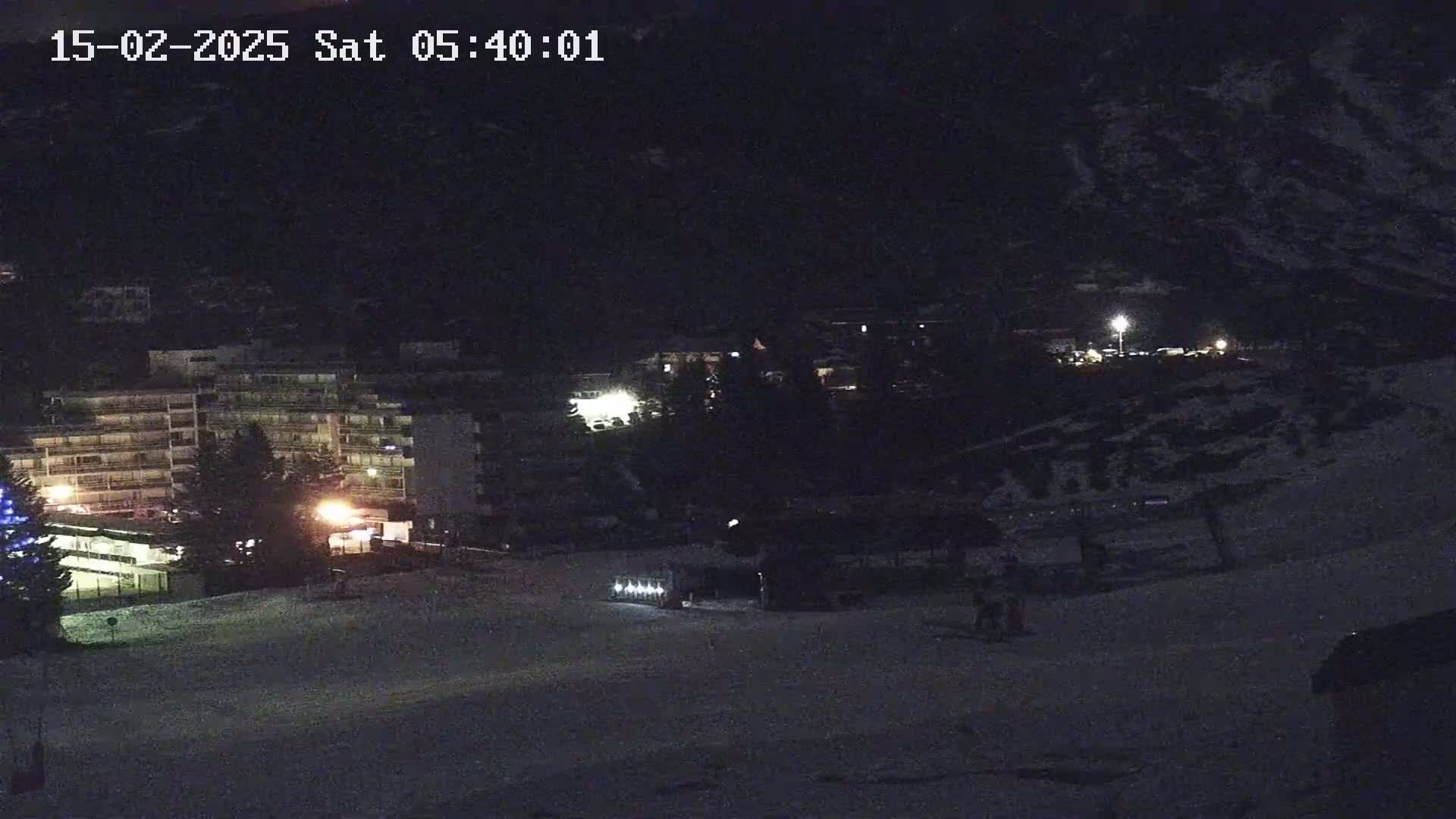 A nighttime, snow-covered landscape shows a multi-story building illuminated with lights, and a sparsely populated area in front of it.