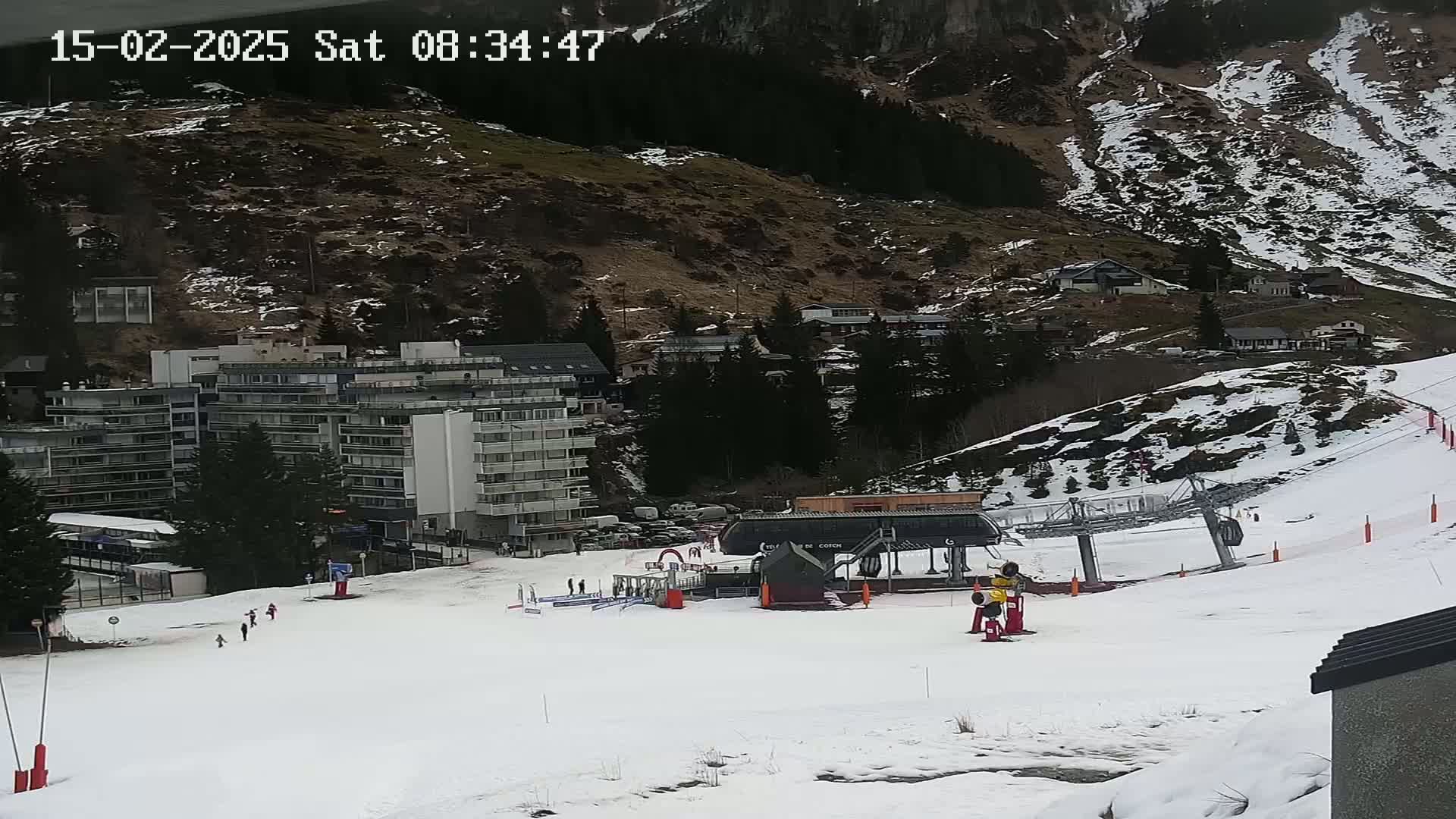 A snow-covered ski slope with a chairlift, a few skiers, and buildings nestled into a mountainside.