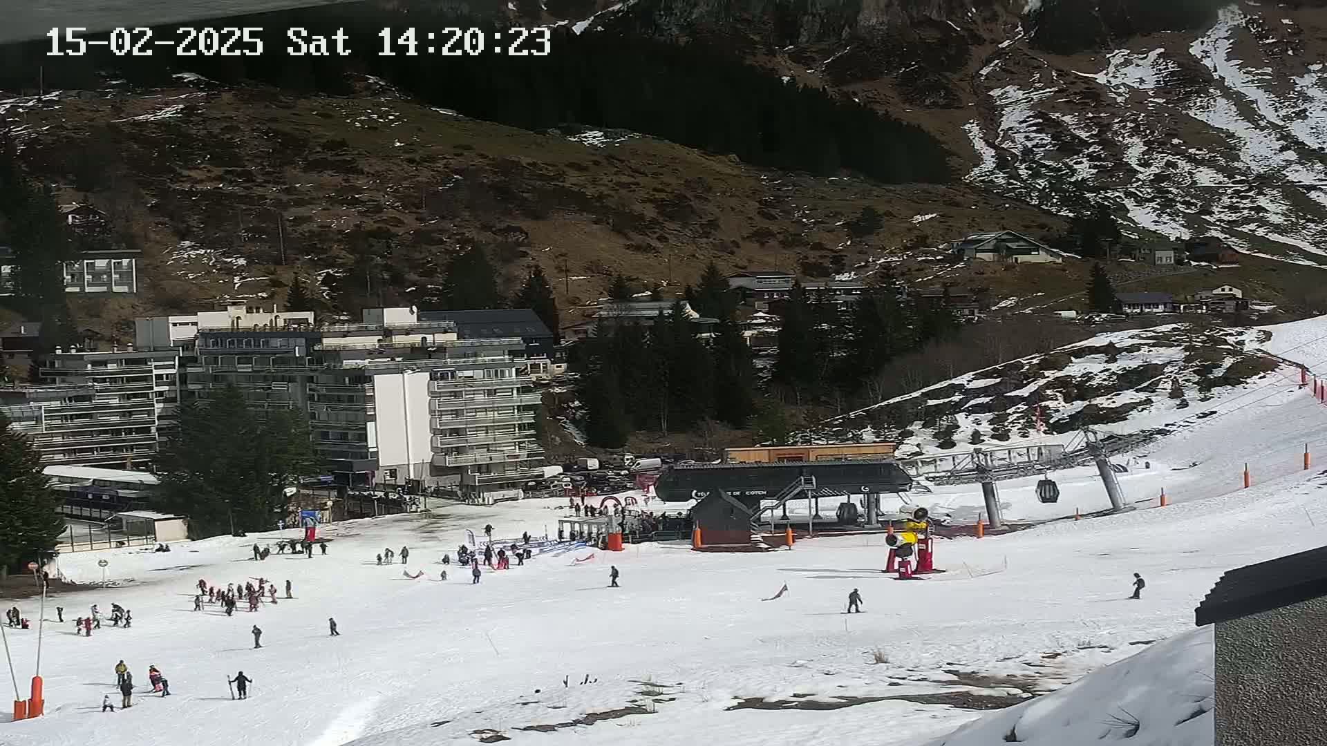 A snowy ski slope with a ski lift and several people skiing and snowboarding, situated below a mountainside with buildings and trees, on a sunny day.