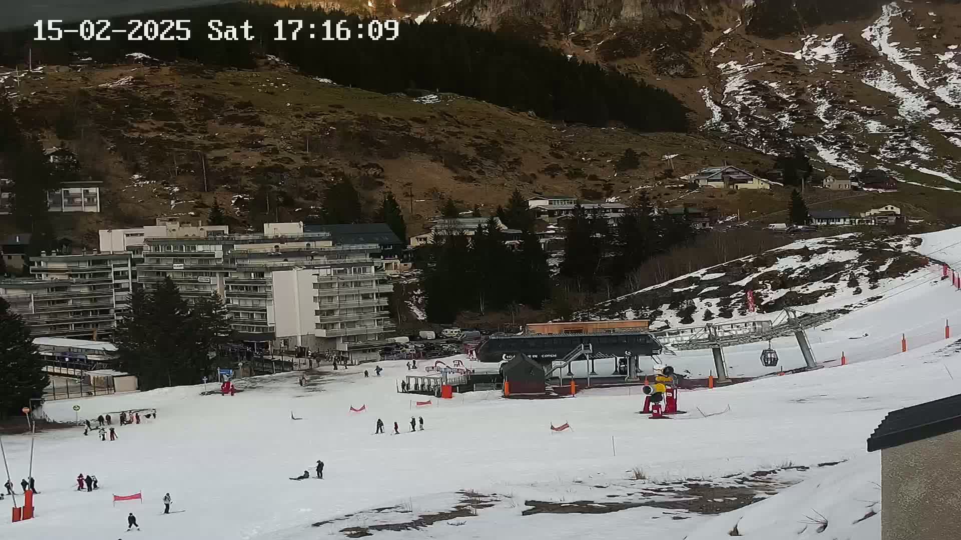 A snowy ski slope with a chairlift and several skiers, situated below a mountainside with buildings and trees.