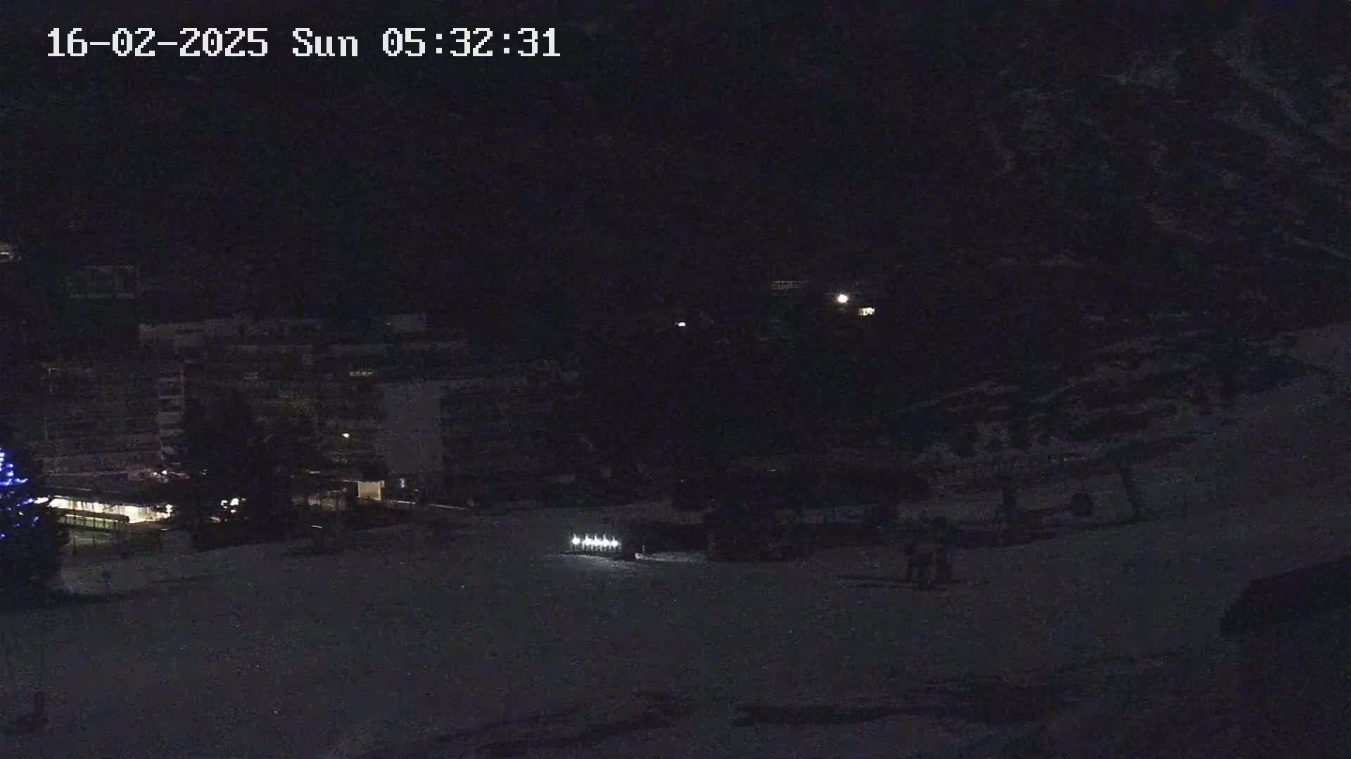 A nighttime, snow-covered landscape shows a row of lights on a slope near a building and a few figures.