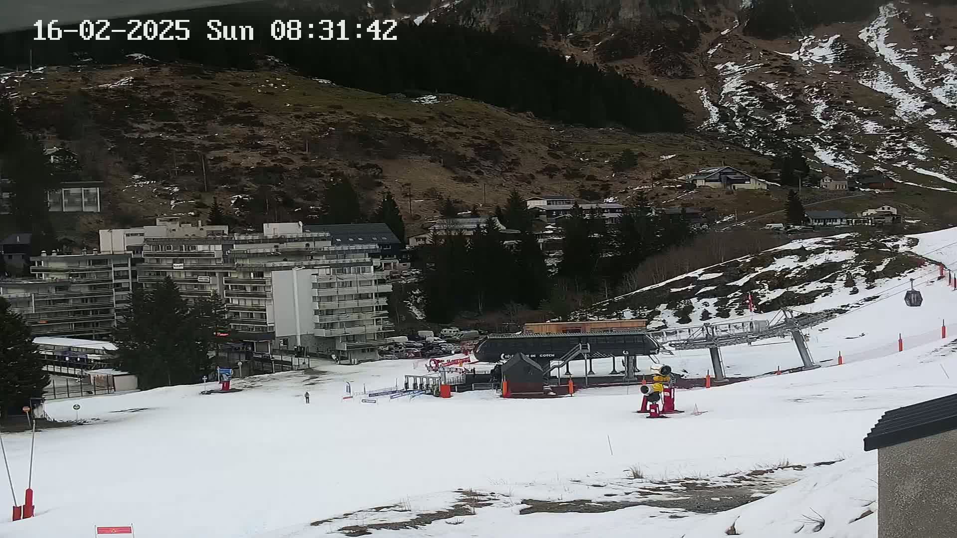 A snow-covered ski slope with a chairlift, a multi-story building, and several smaller buildings nestled against a mountainside on a cloudy day.