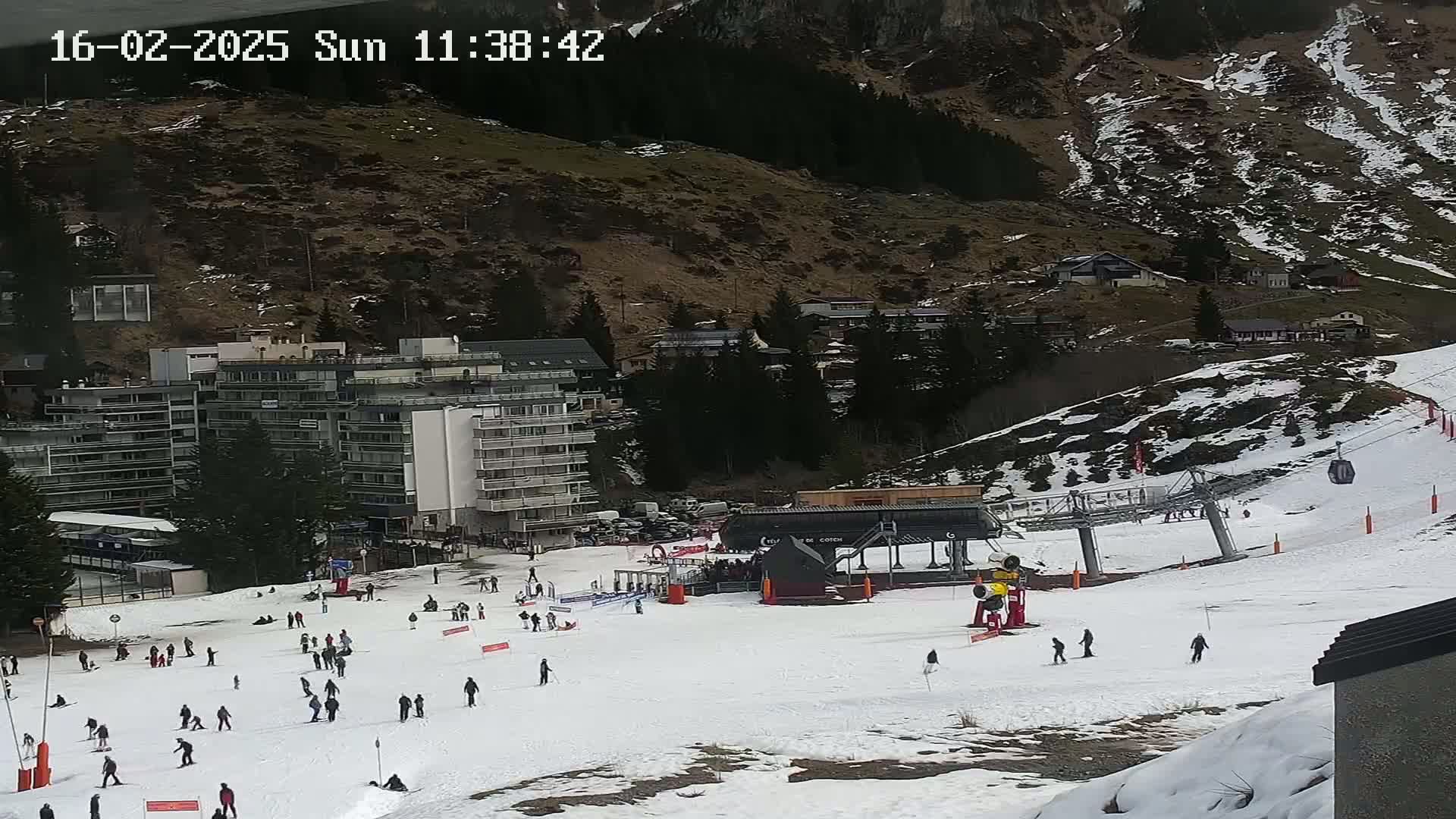 A ski slope with several people skiing and snowboarding, a ski lift, and buildings in the background on a cloudy day.