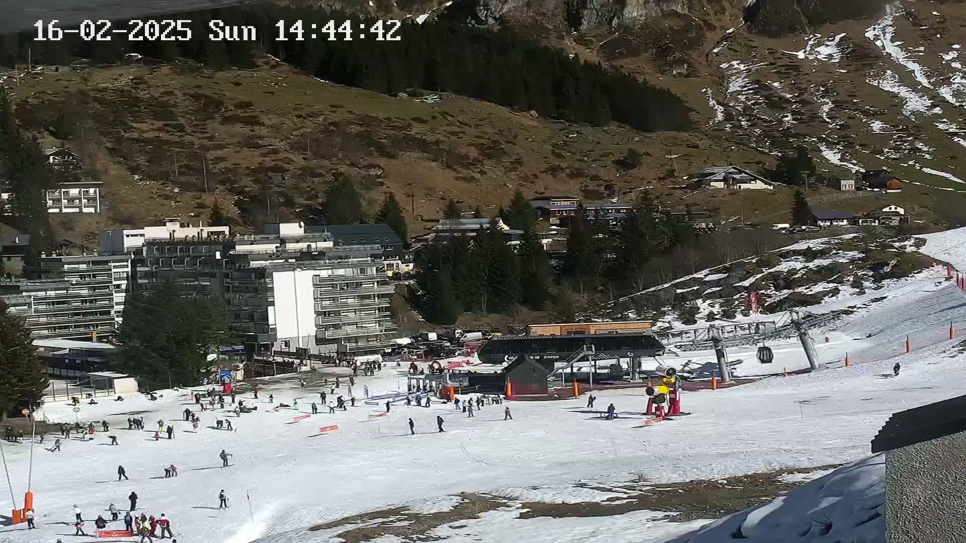 A sunny ski slope with scattered skiers and snowboarders near a ski lift and several buildings on a mountainside.
