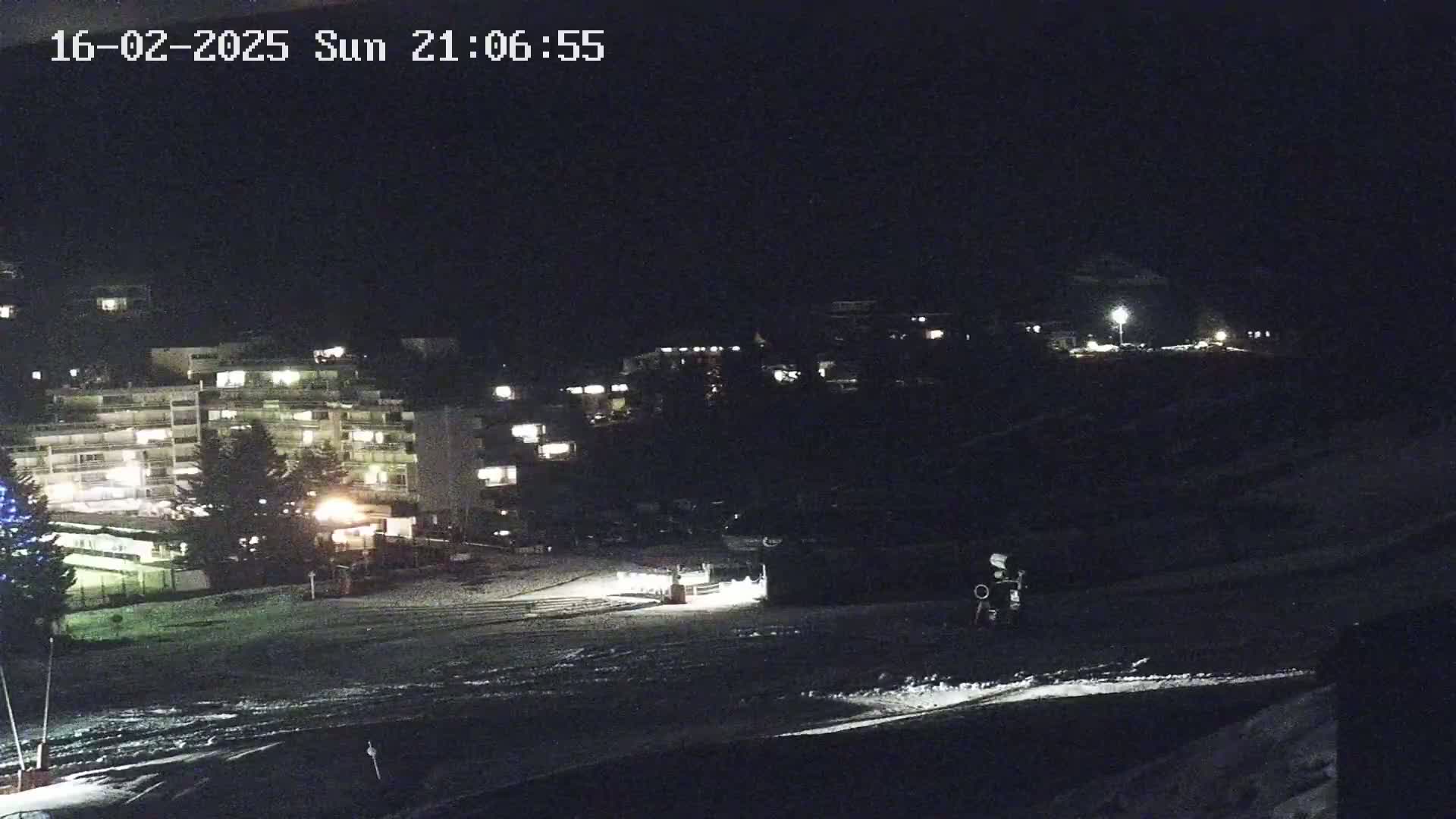 A nighttime view of a snow-covered area with illuminated buildings in the background under a dark sky.