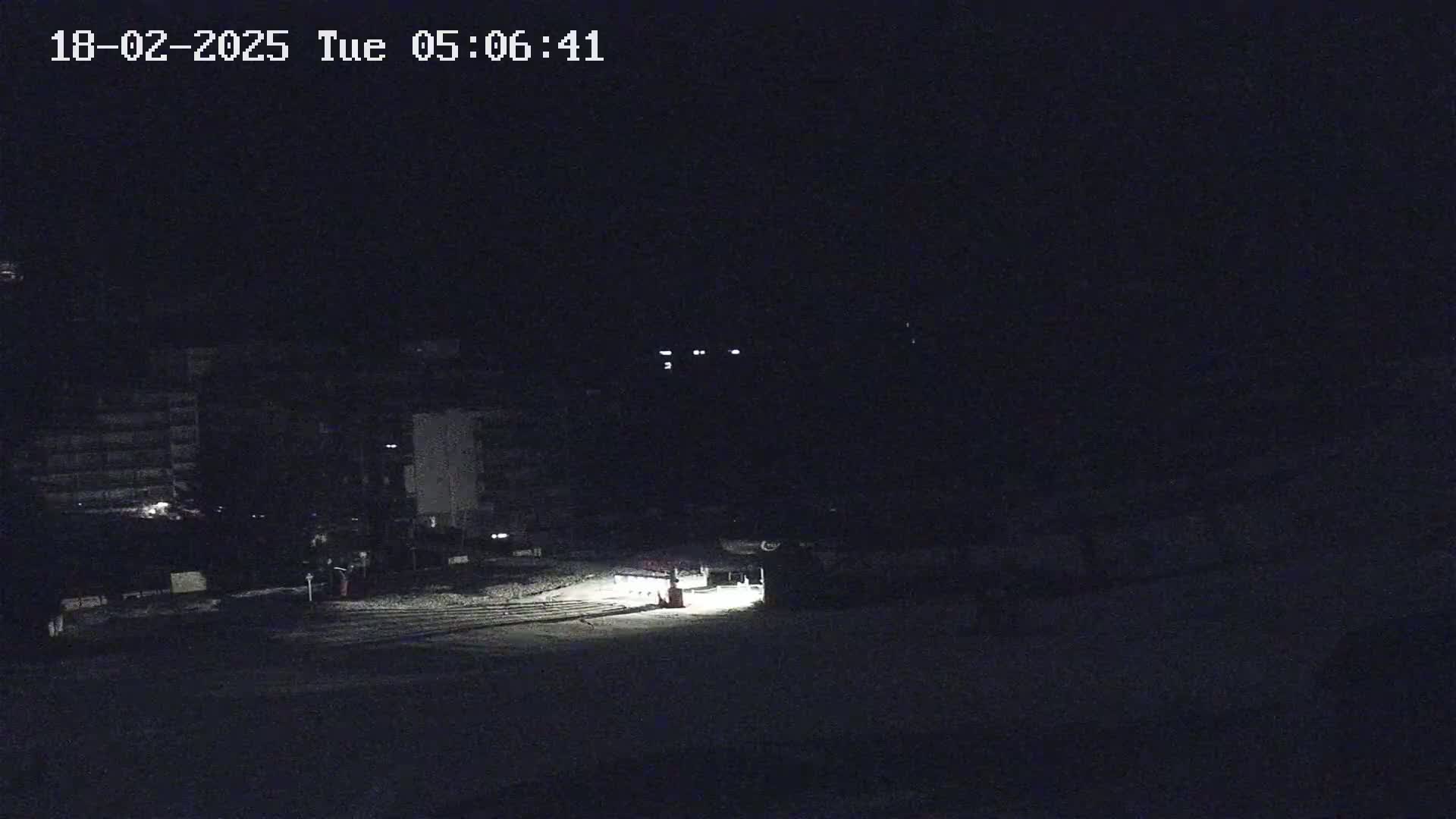 A nighttime, snow-covered scene shows dimly lit buildings and a vehicle with headlights illuminating a small area in the foreground.