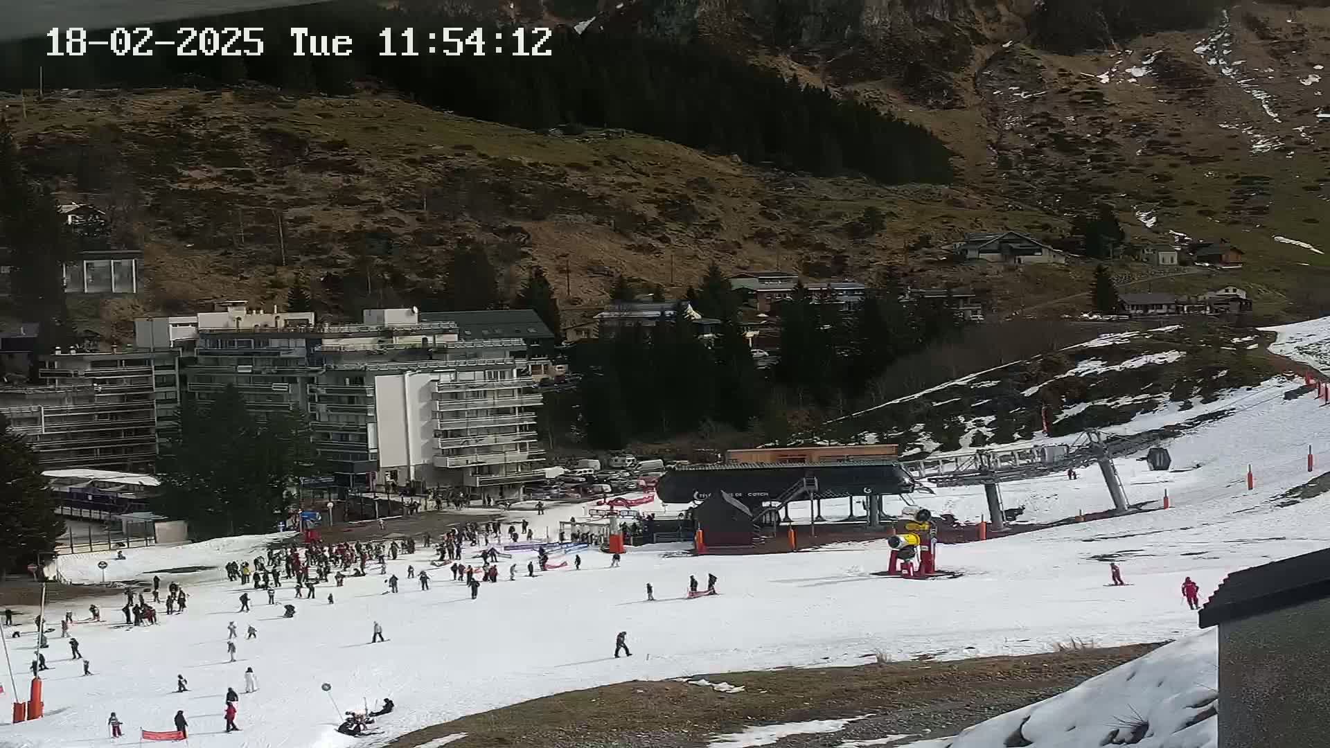 A ski slope with many people skiing and snowboarding, a ski lift, several buildings, and a mountainside with patchy snow cover under a partly cloudy sky.