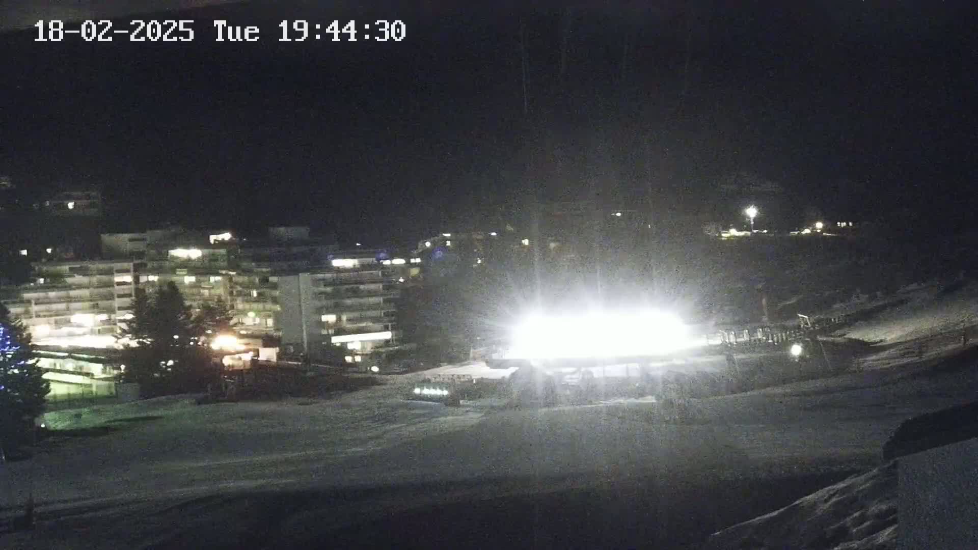 A brightly lit snow-covered slope at night, with a multi-story building illuminated in the background.