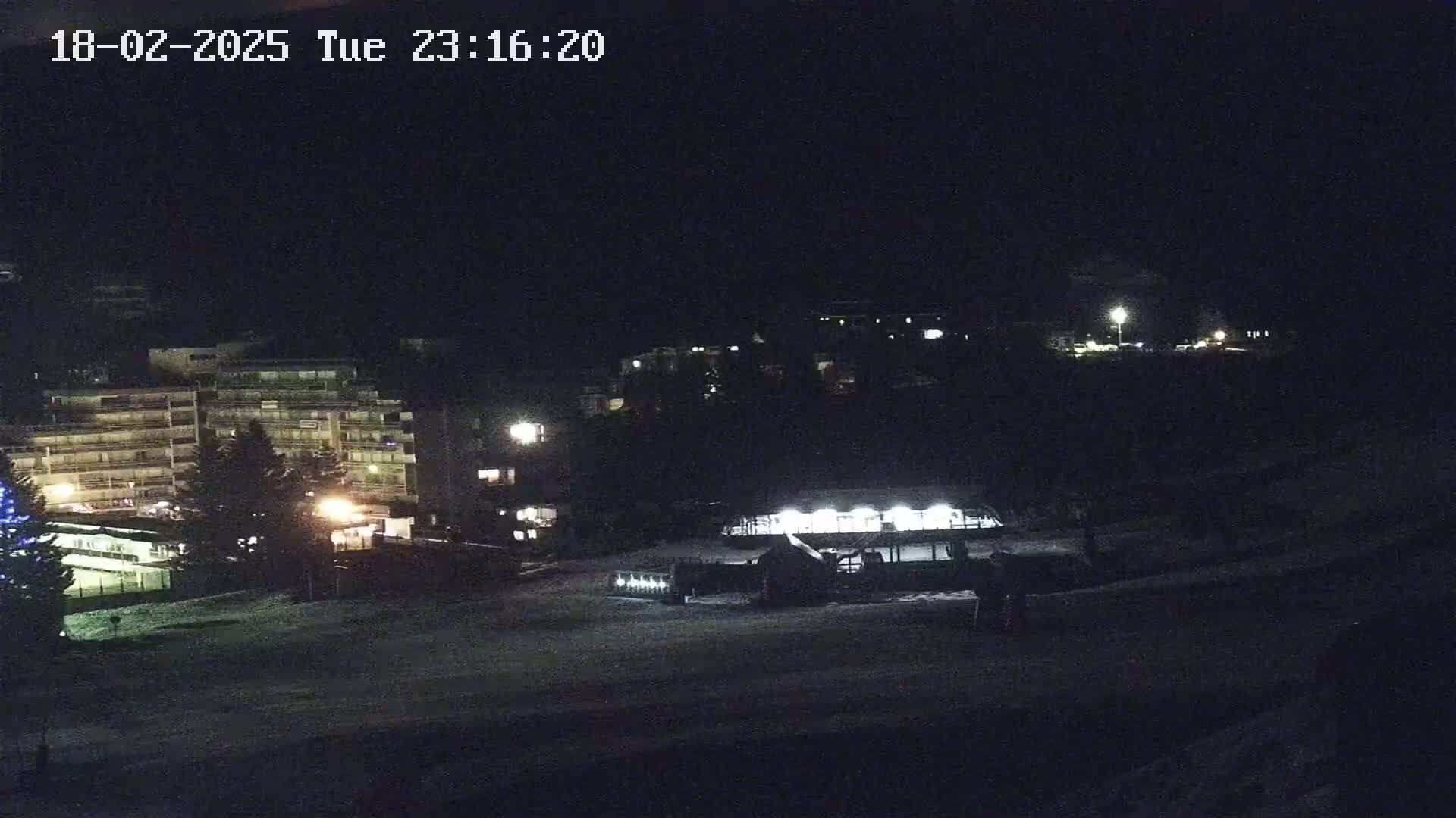A nighttime view of snow-covered ground with buildings and lights visible in the distance under a dark sky.