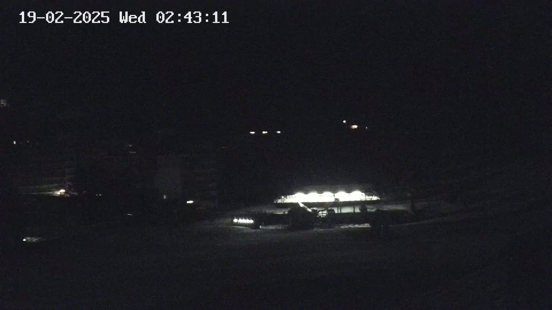 A snowplow is illuminated at night in a snowy area with dimly lit buildings and vehicles in the background.