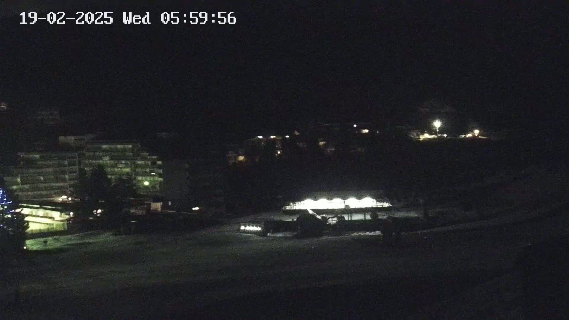 A nighttime, snow-covered scene shows a building complex, a snowcat, and a few sparsely lit structures in the distance under a dark sky.
