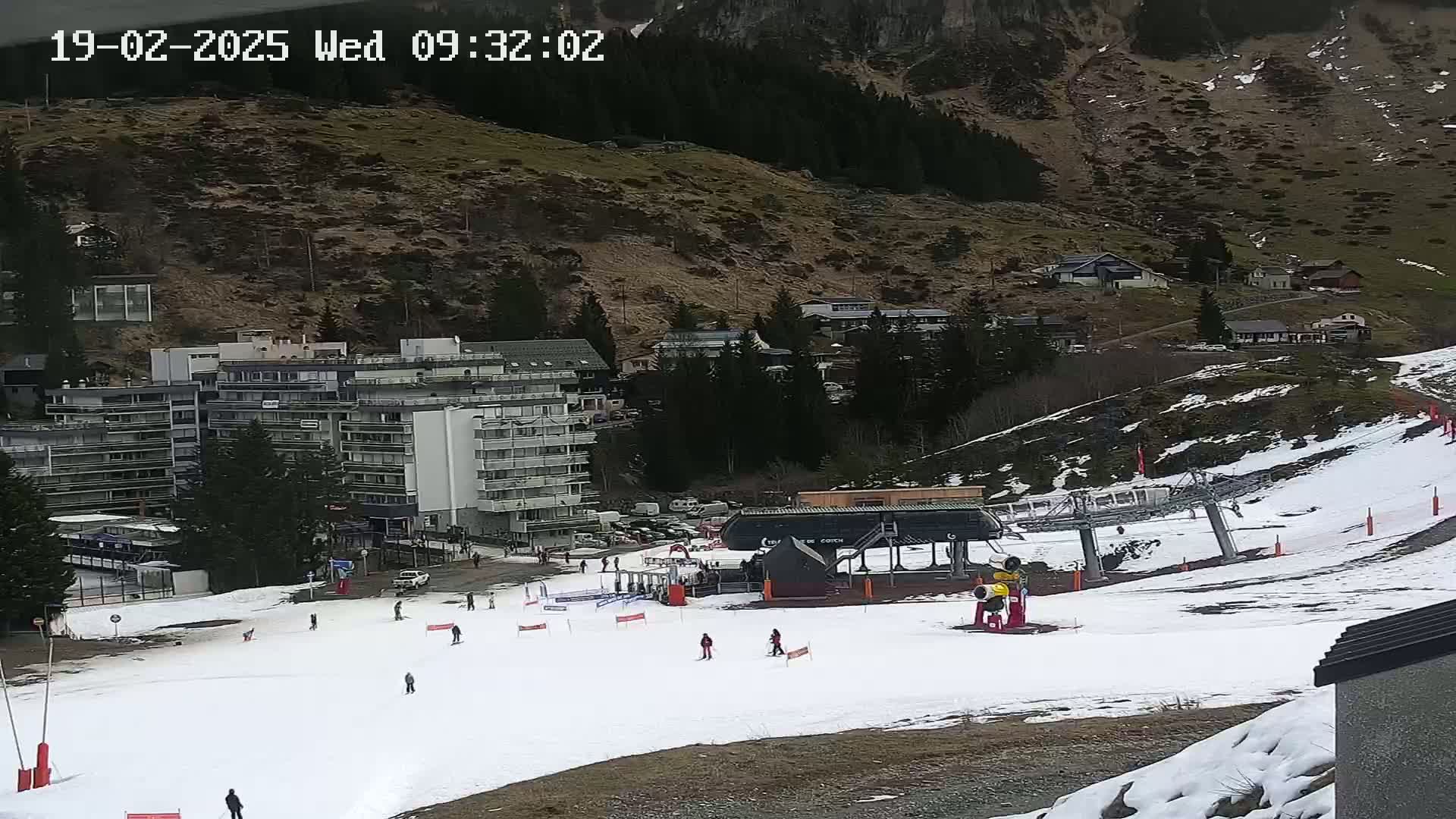 A ski slope with a chairlift and several skiers, situated near a large building and a mountainside village, under a mostly cloudy sky.