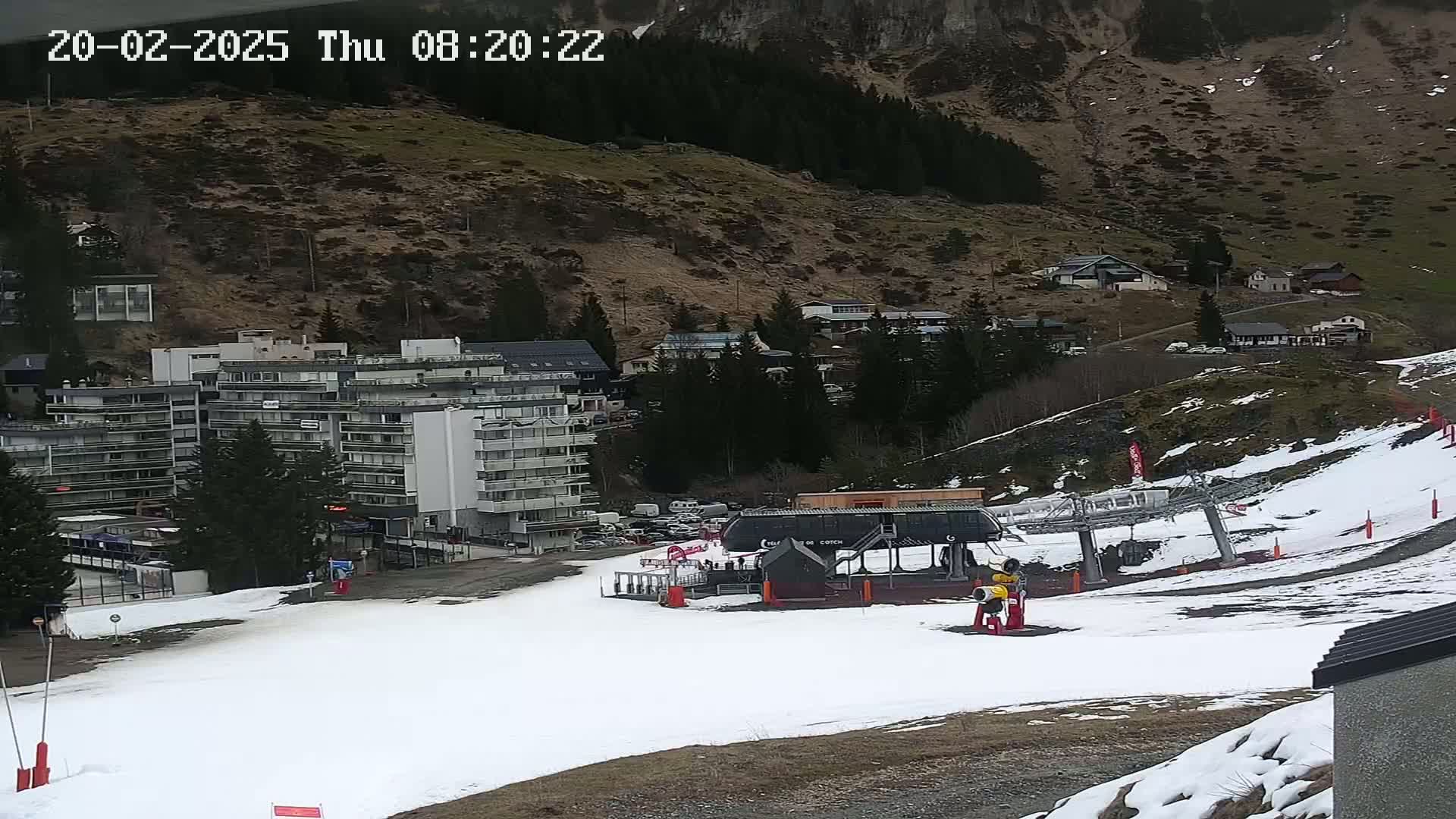 A ski lift base station sits at the bottom of a snow-covered slope near a multi-story building and a village nestled in a mountainside, under an overcast sky.