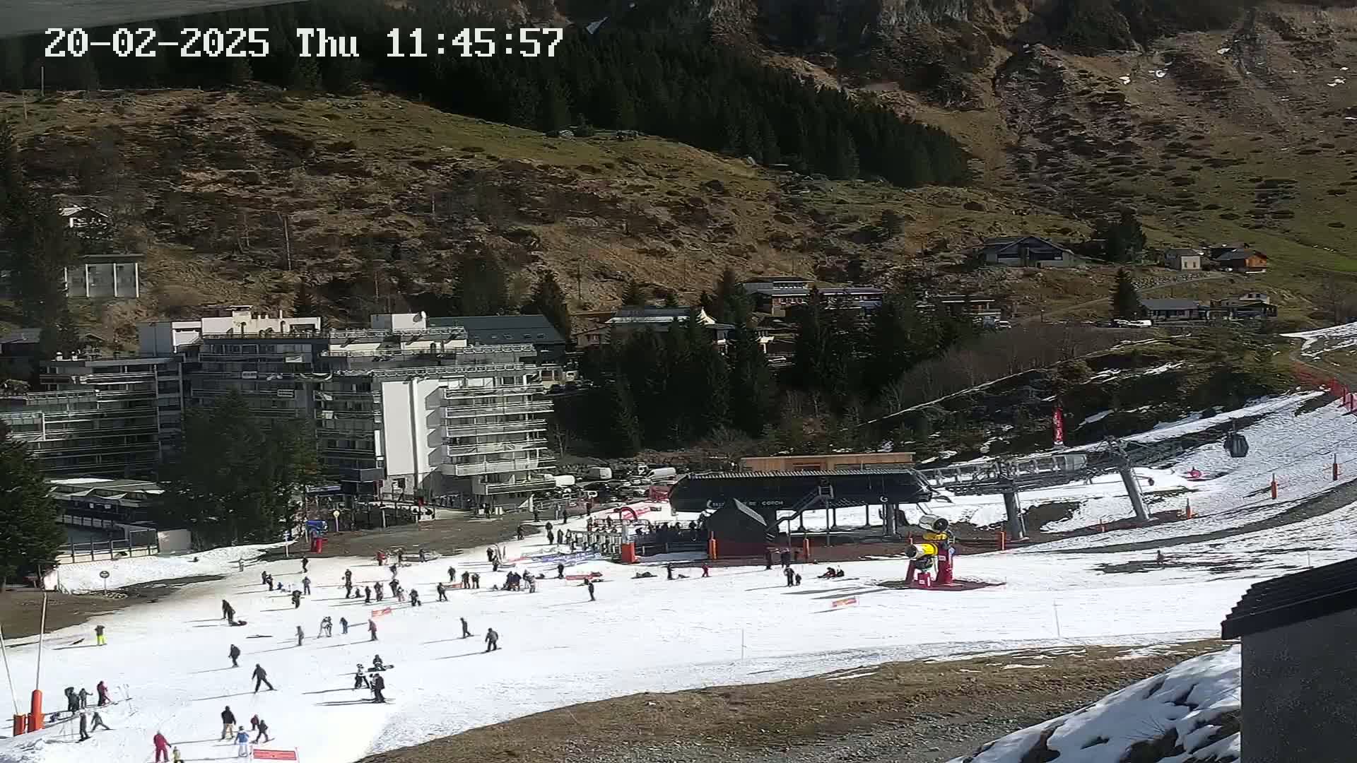 A snowy ski slope with several skiers and snowboarders, a chairlift, and buildings nestled in a mountainside on a sunny day.