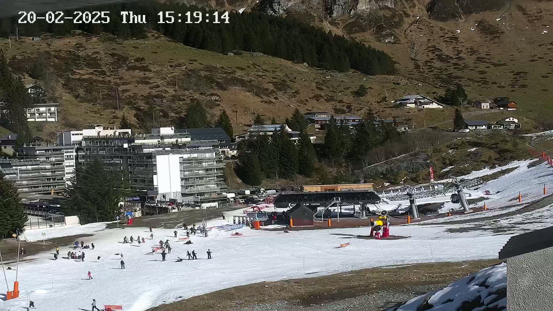 A ski slope with a chairlift and several people skiing and snowboarding on a sunny day, with buildings and a mountain in the background.