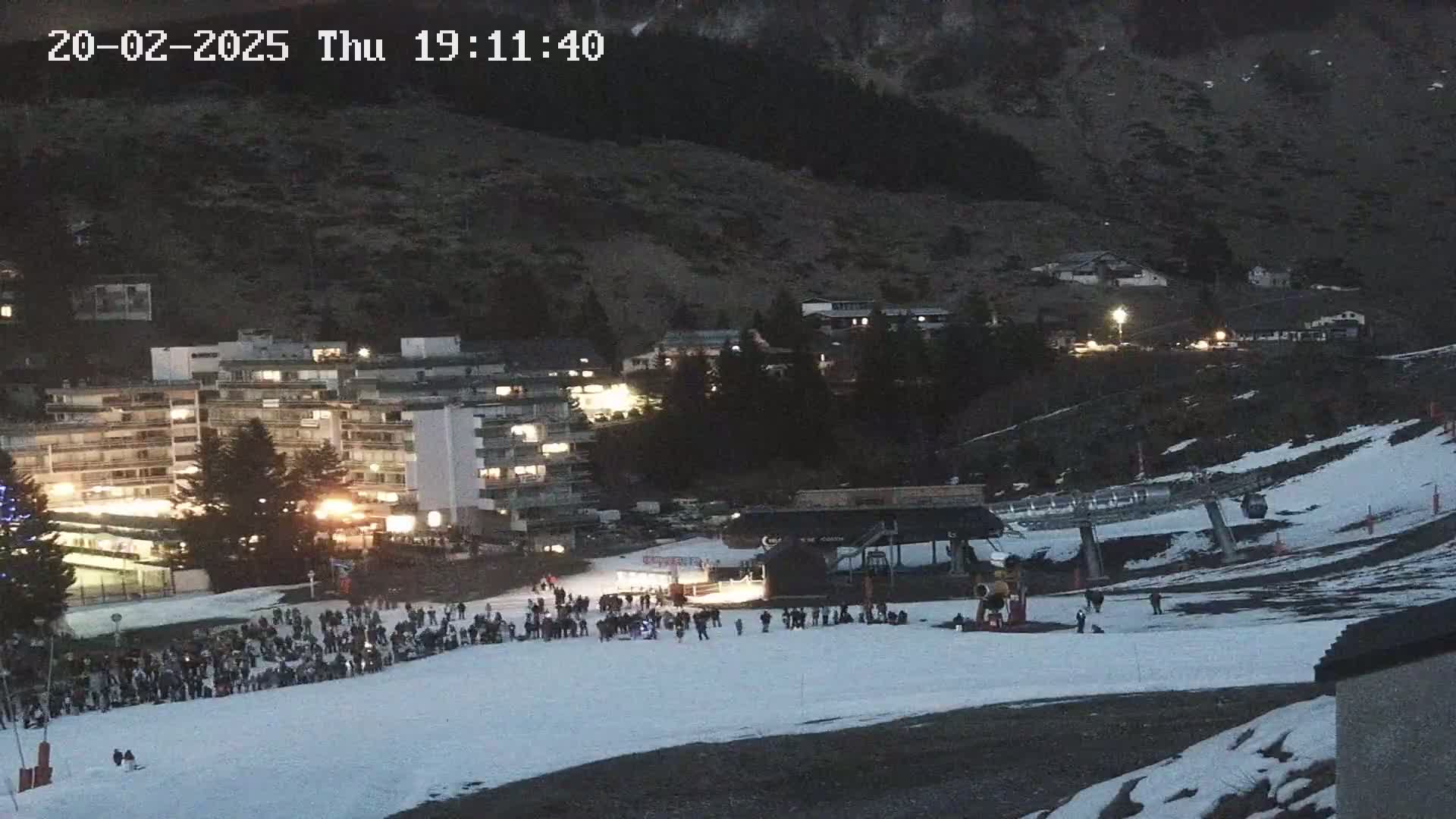 A snowy ski slope at night, with a crowd of people gathered near a ski lift and buildings with lights on in the background under a dark sky.