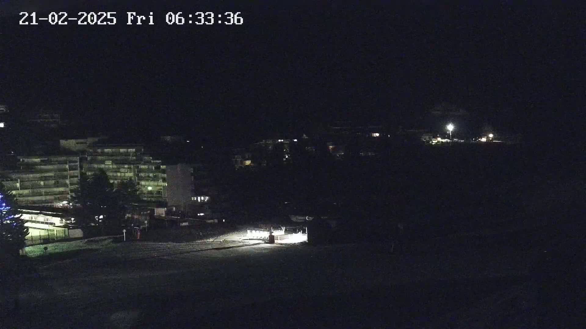 A dark, nighttime view of a snow-covered area with buildings and a few lights visible in the distance.