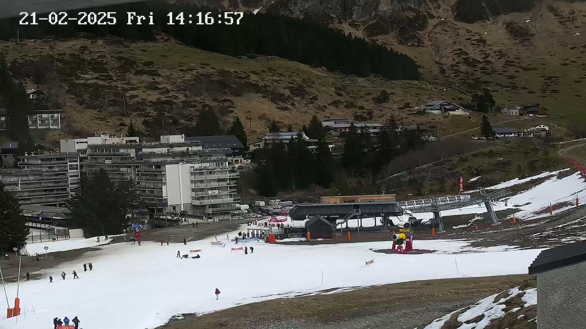 A ski resort with a chairlift, several buildings, and a sparsely snow-covered slope is visible on a partly cloudy day.