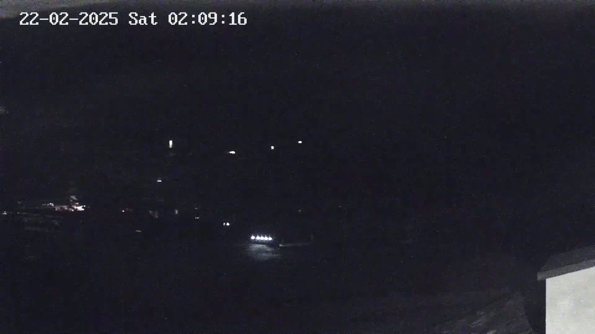 A nighttime, dark scene shows a few distant lights and a vehicle's headlights on a road.